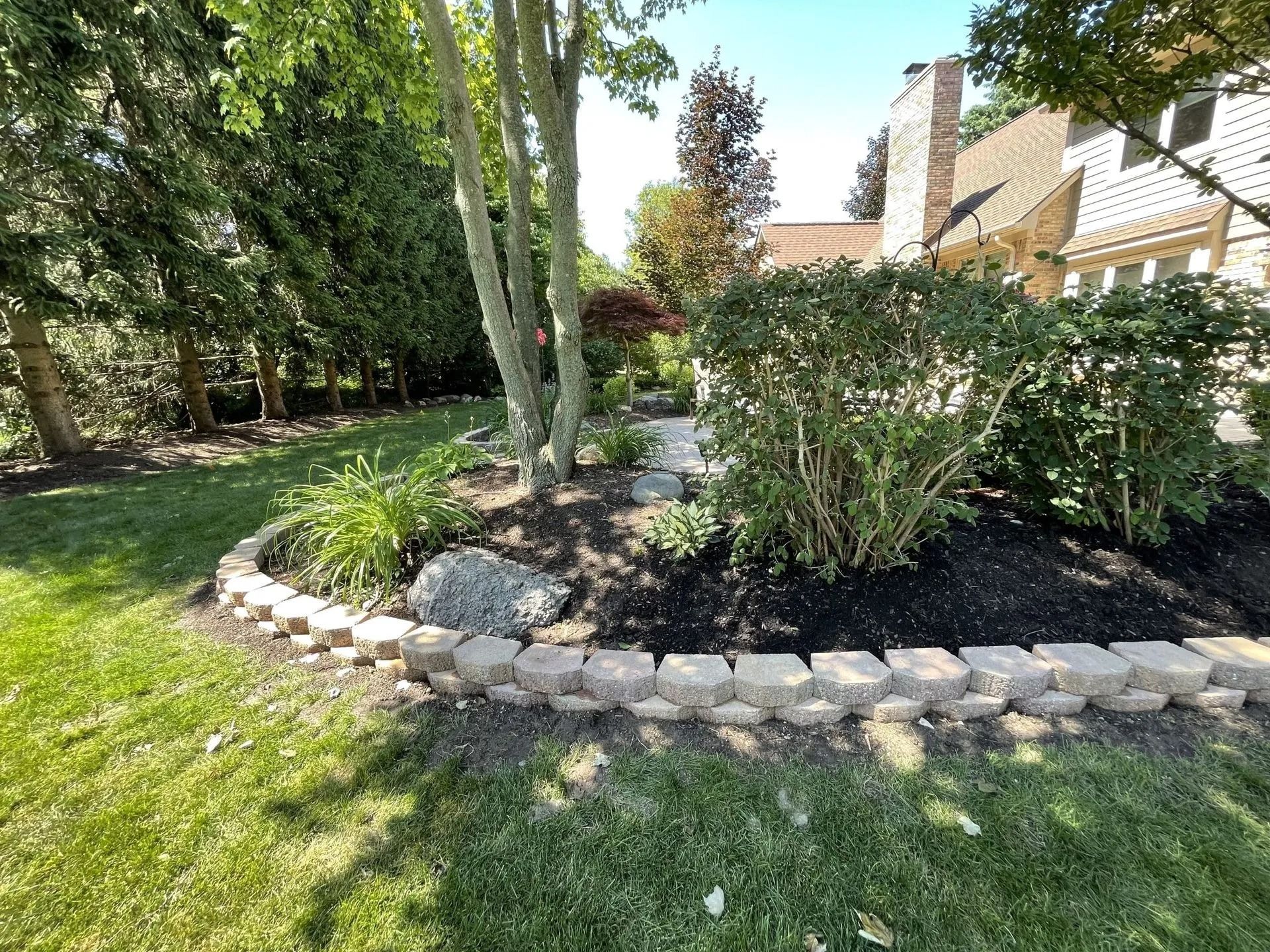Landscaped front yard with a stone-edged flower bed, mulch, shrubs, trees, and a house in the background.