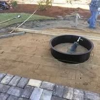 Fire pit ring set in leveled sand, near a brick patio, person in background.