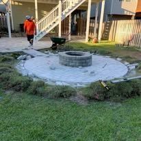 Man stands near a circular stone patio with a fire pit, next to grass.