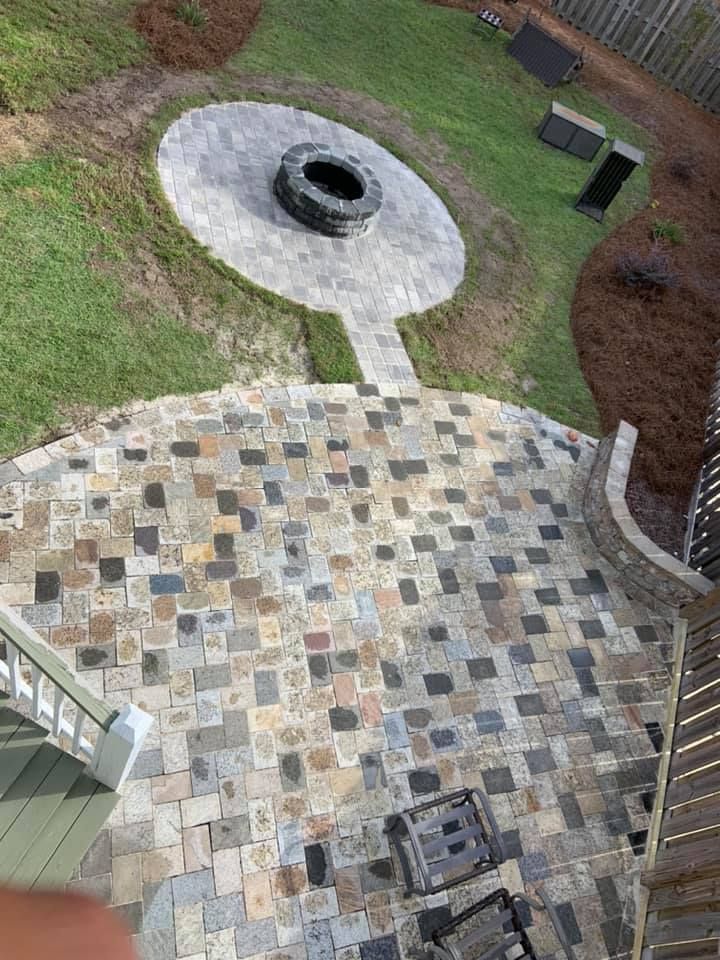 Overhead view of a stone patio, steps, walkway to a circular fire pit area in a backyard. Green grass surrounds.