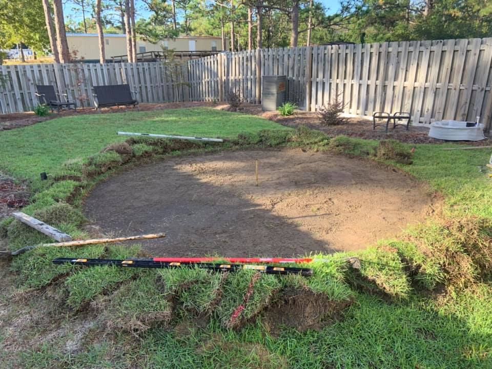 A backyard with a circular area dug out of the grass. Grass sod borders the dirt circle. A wooden fence is in the background.