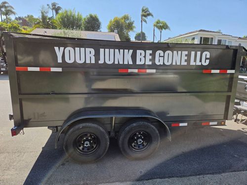 Black dump trailer with “YOUR JUNK BE GONE LLC” sign parked outdoors in sunlight