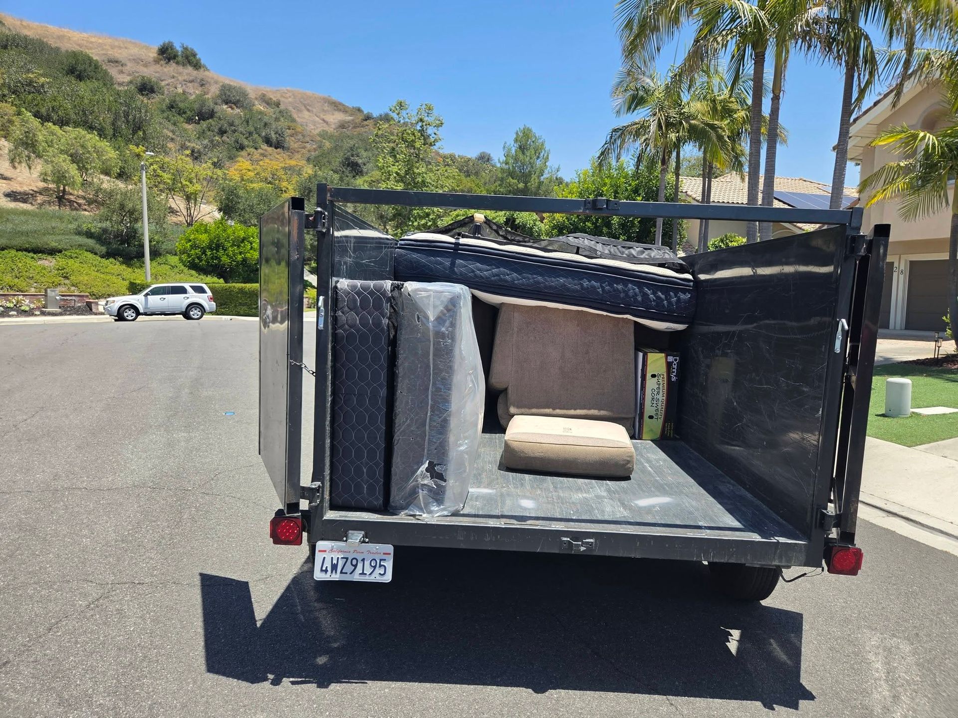 Small black utility trailer with an open cargo area parked on a suburban street near palm trees and hills.