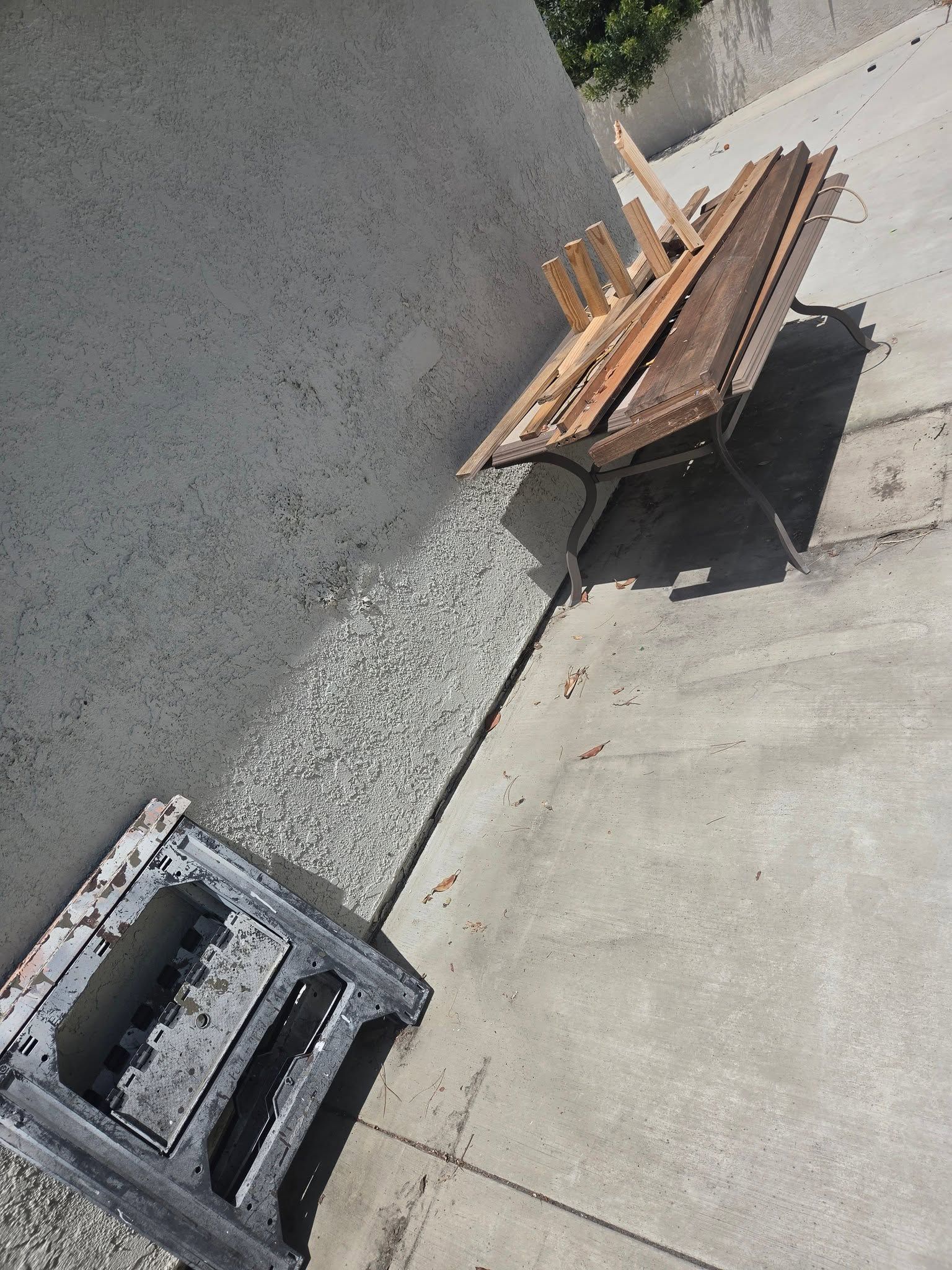 Pallets and lumber stacked beside a concrete wall on a sidewalk or driveway, with a cinder block in the foreground