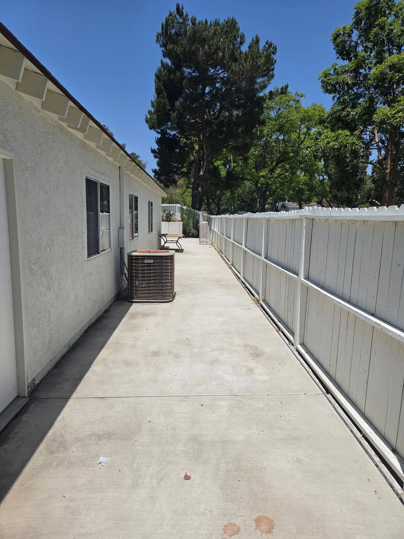 Narrow concrete side yard beside a white house and fence, with a crate near the wall and trees at the end.