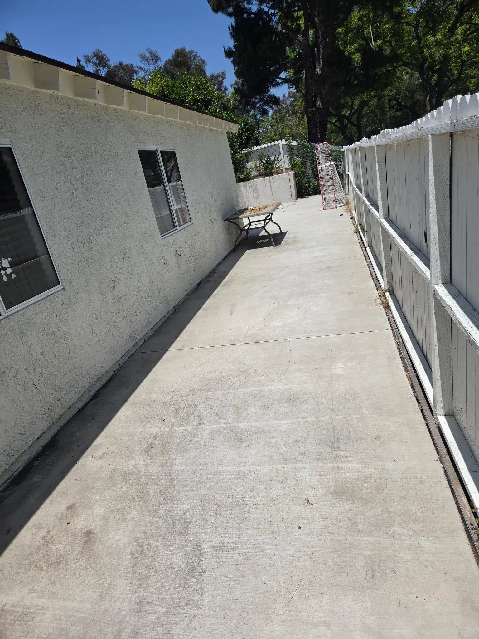 Concrete side yard beside a white house and fence, with a wheelbarrow near the end.