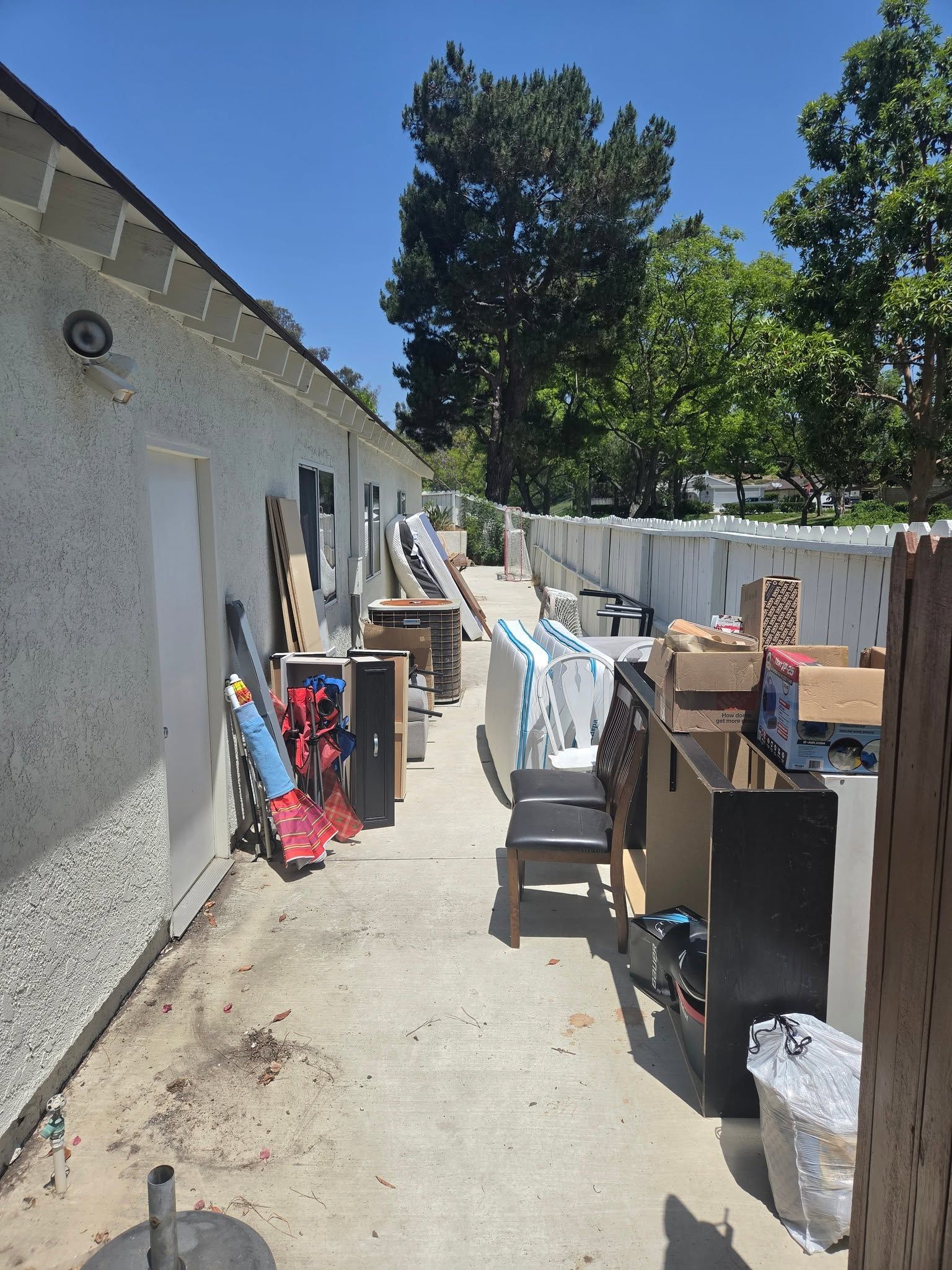 Narrow outdoor alley beside a stucco building, cluttered with stacked household items and bins in sunlight.