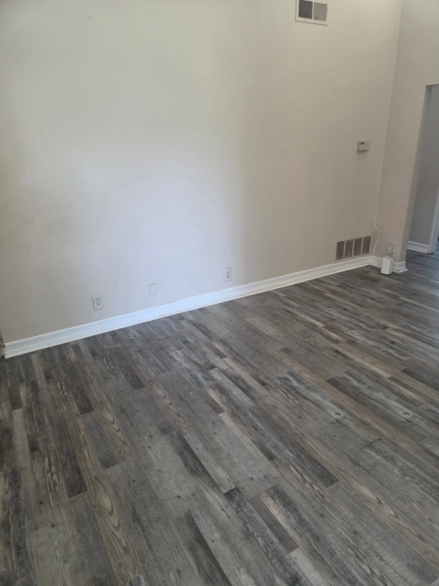 Empty room with gray wood-look flooring and white walls, showing a small wall opening and outlets.