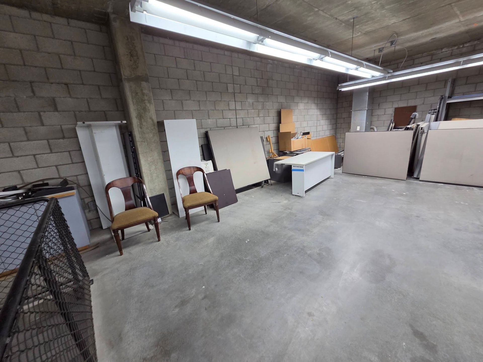 Empty workshop with cinderblock walls, concrete floor, chairs, desks, and stacked boards under fluorescent lights