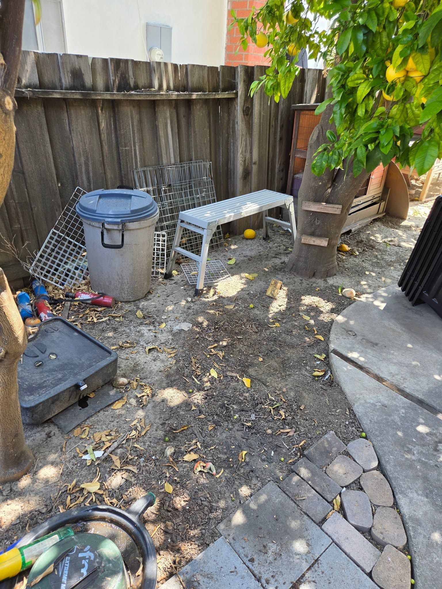 Backyard patio with trash bins, bench, gravel ground, and wooden fence under a tree