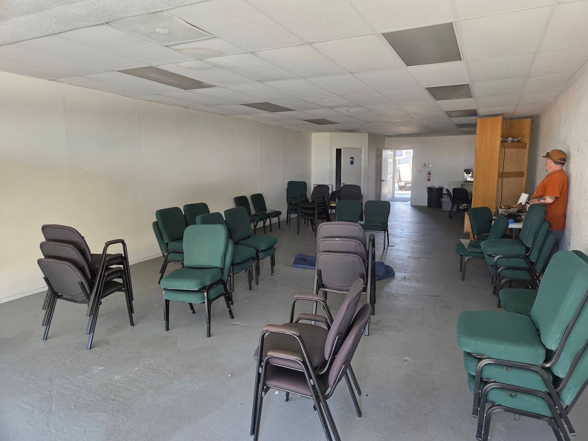 Rows of green and gray chairs in an empty waiting room with a person standing near the back wall.