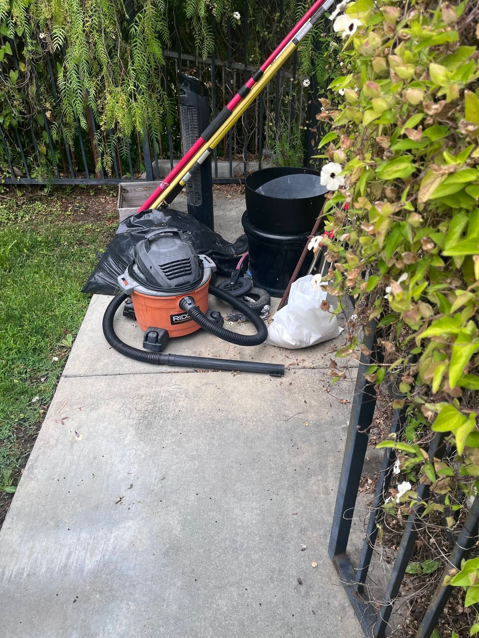 Leaf blower, rake, and trash bag on a sidewalk beside a hedge and fence.
