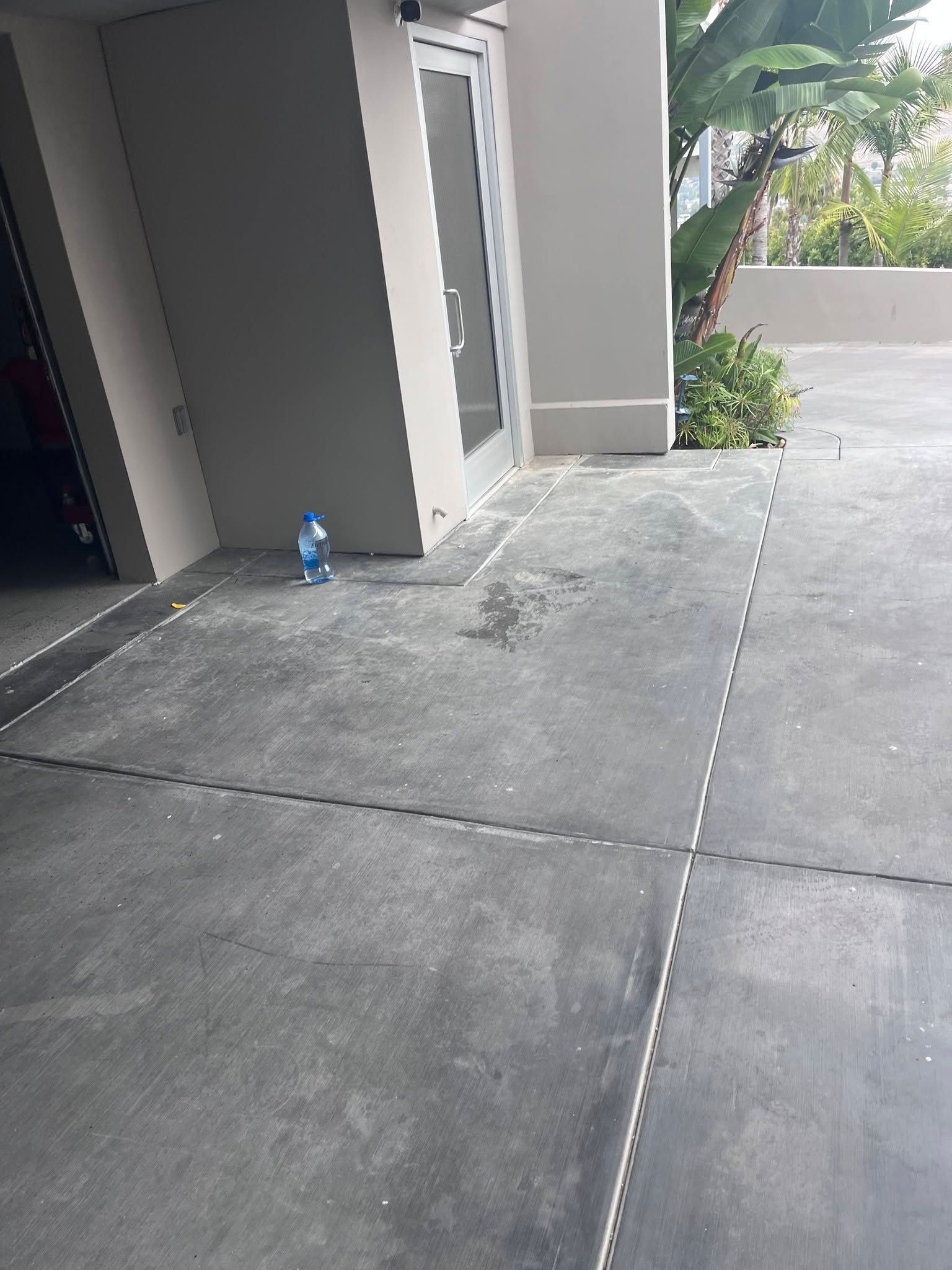 Concrete walkway beside a gray building, with a blue water bottle near the wall and trees ahead.