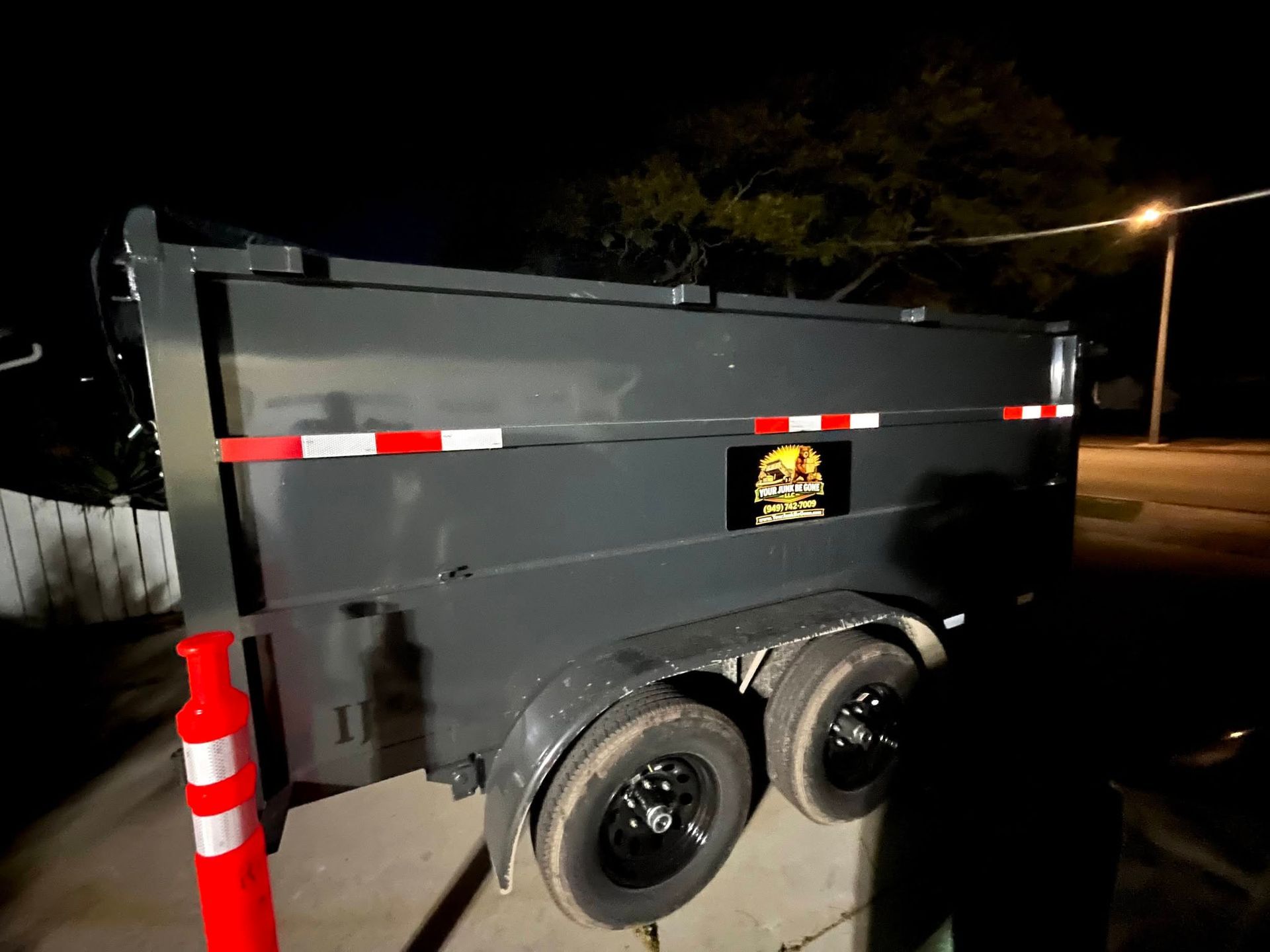 Black utility trailer with two wheels parked at night beside a streetlight and orange cone.