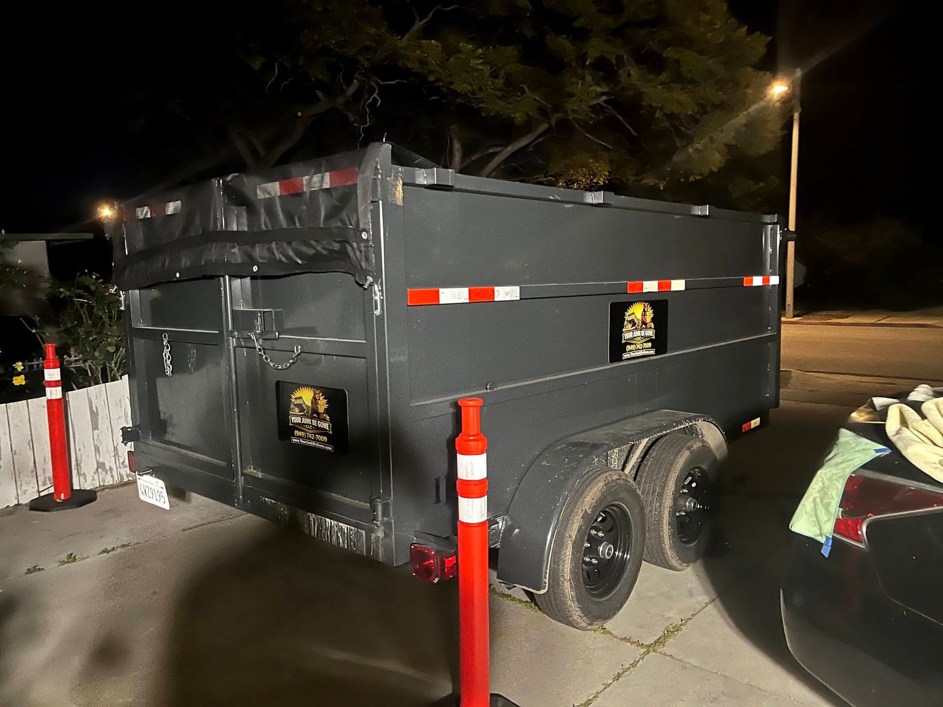 Black dump trailer parked at night beside a car, with orange traffic cones and reflective stripes.
