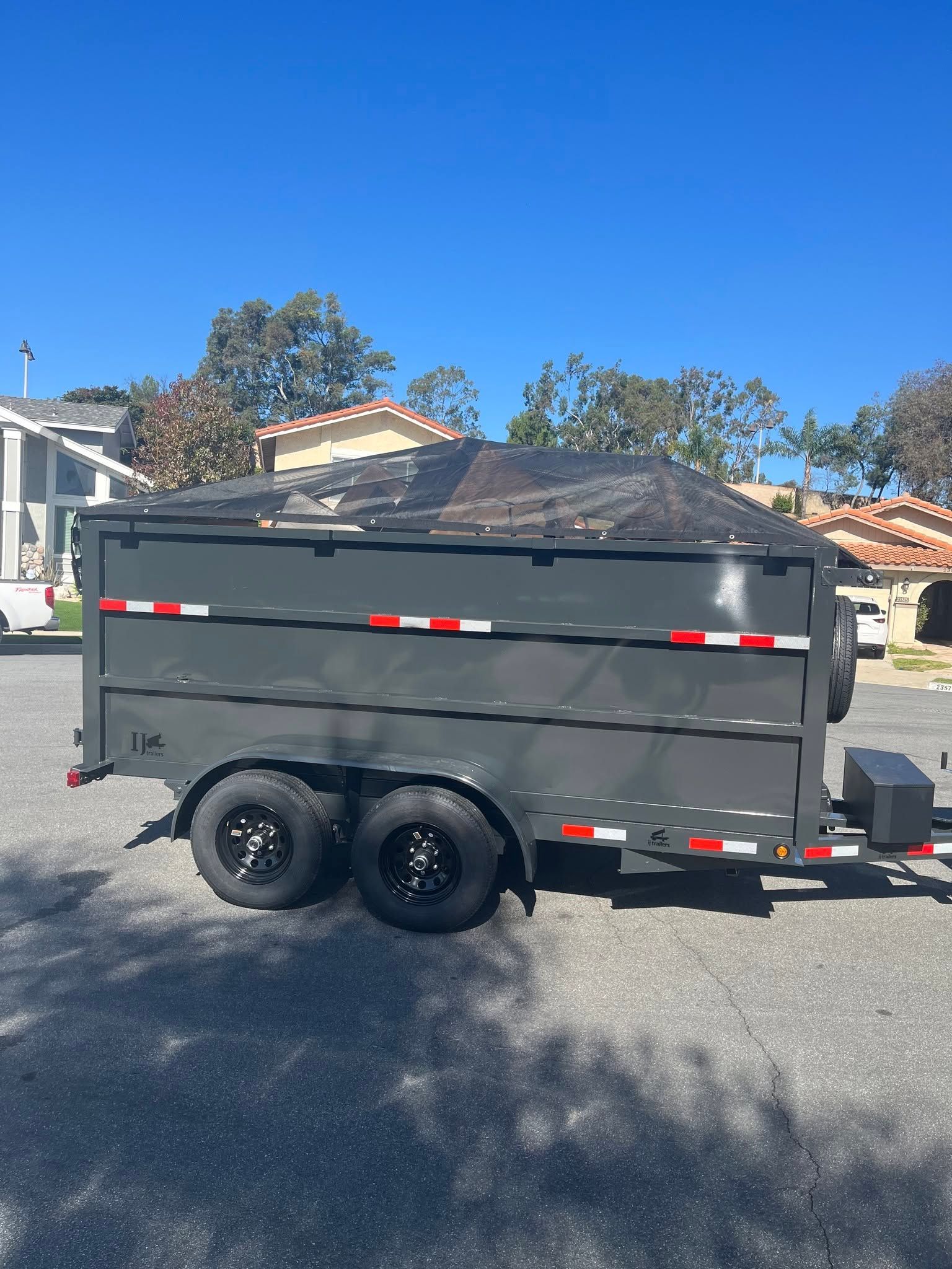 Gray dual-axle dump trailer parked on a residential street in bright sunlight
