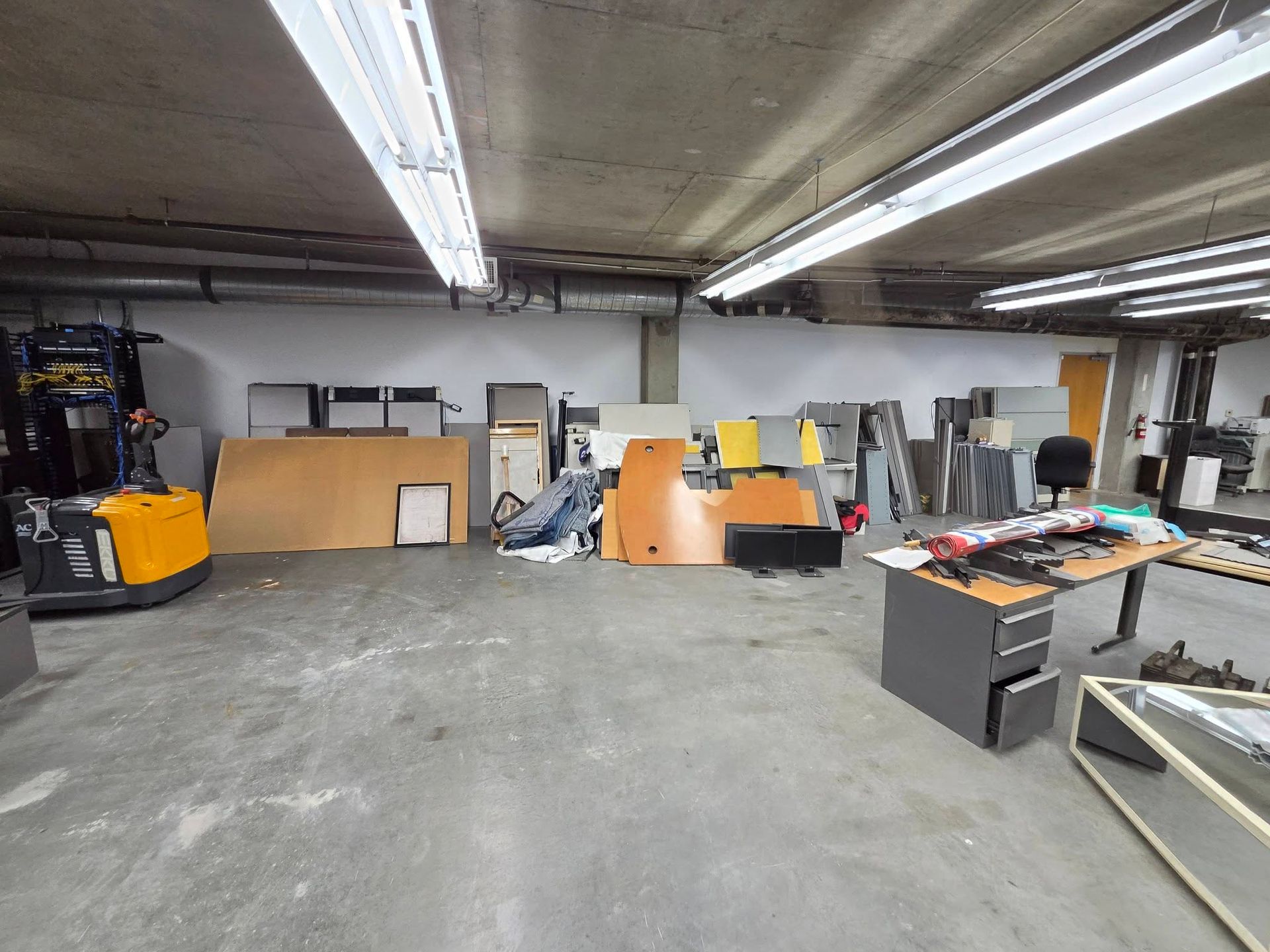 Wide garage workshop with concrete floor, fluorescent lights, shelves, tools, and stacked materials.