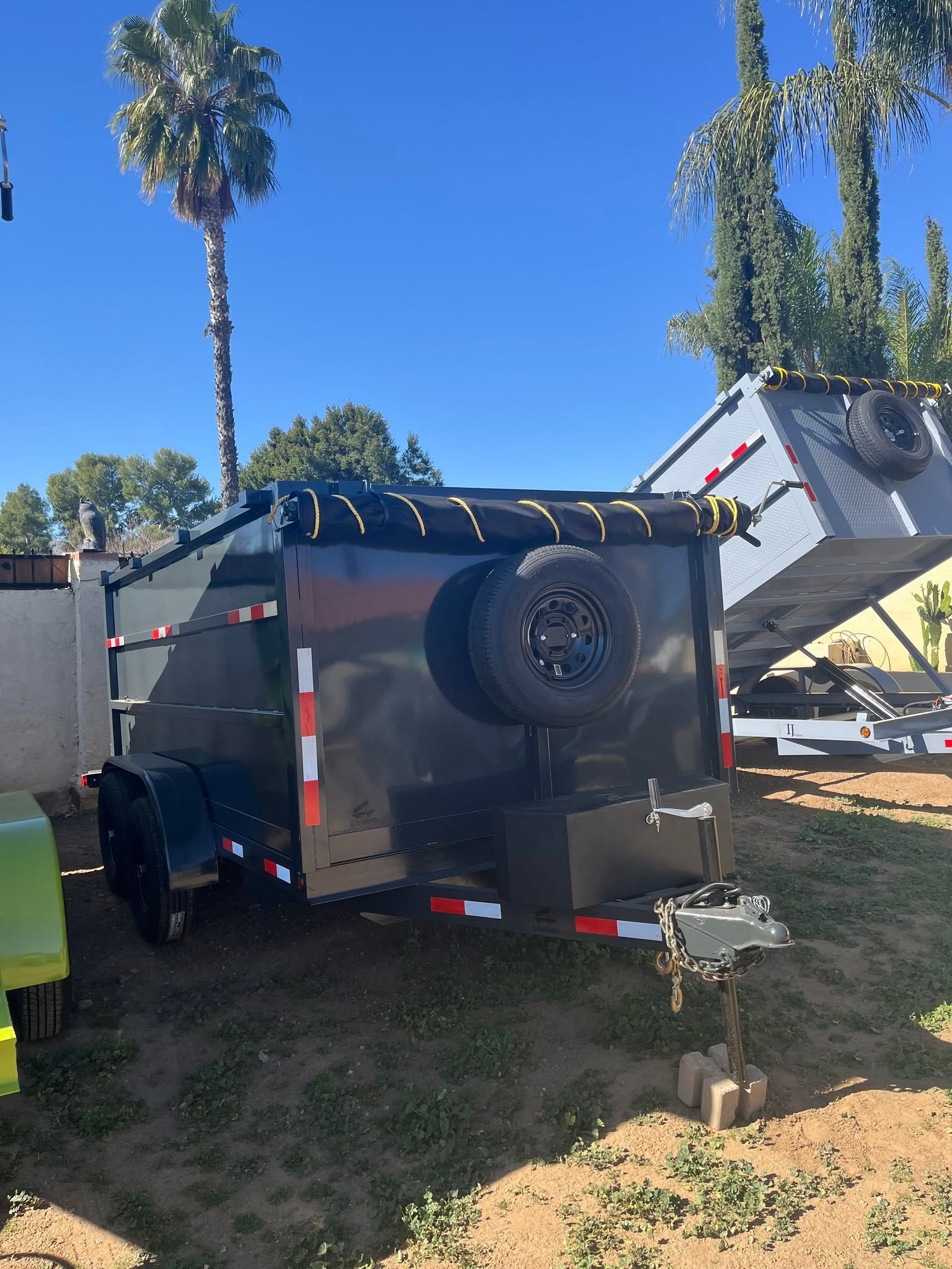 Black off-road trailer with spare tire parked in a sunny yard beside a small ramp and palm trees.