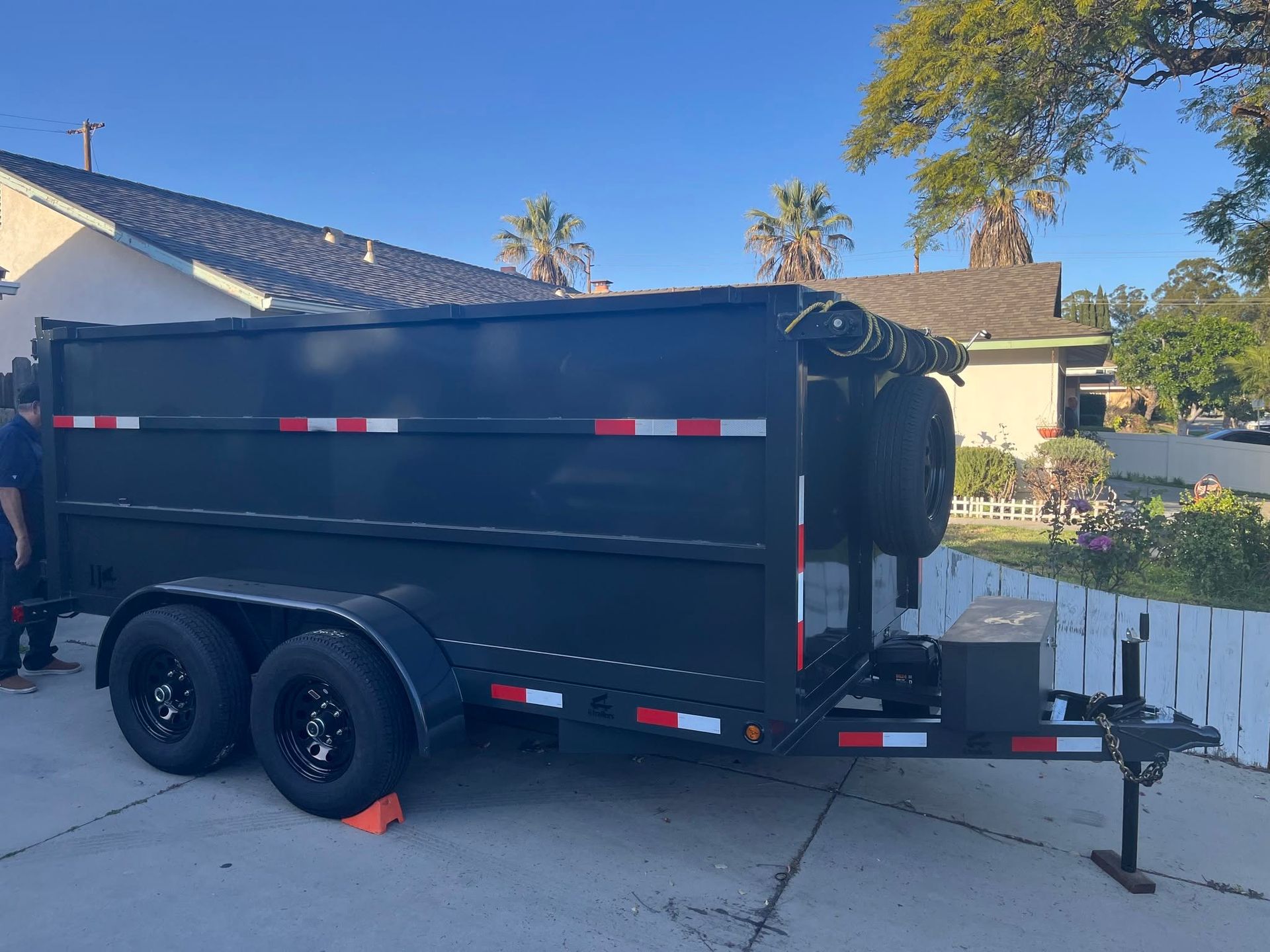 Black enclosed dual-axle trailer parked in a driveway beside a house and white fence