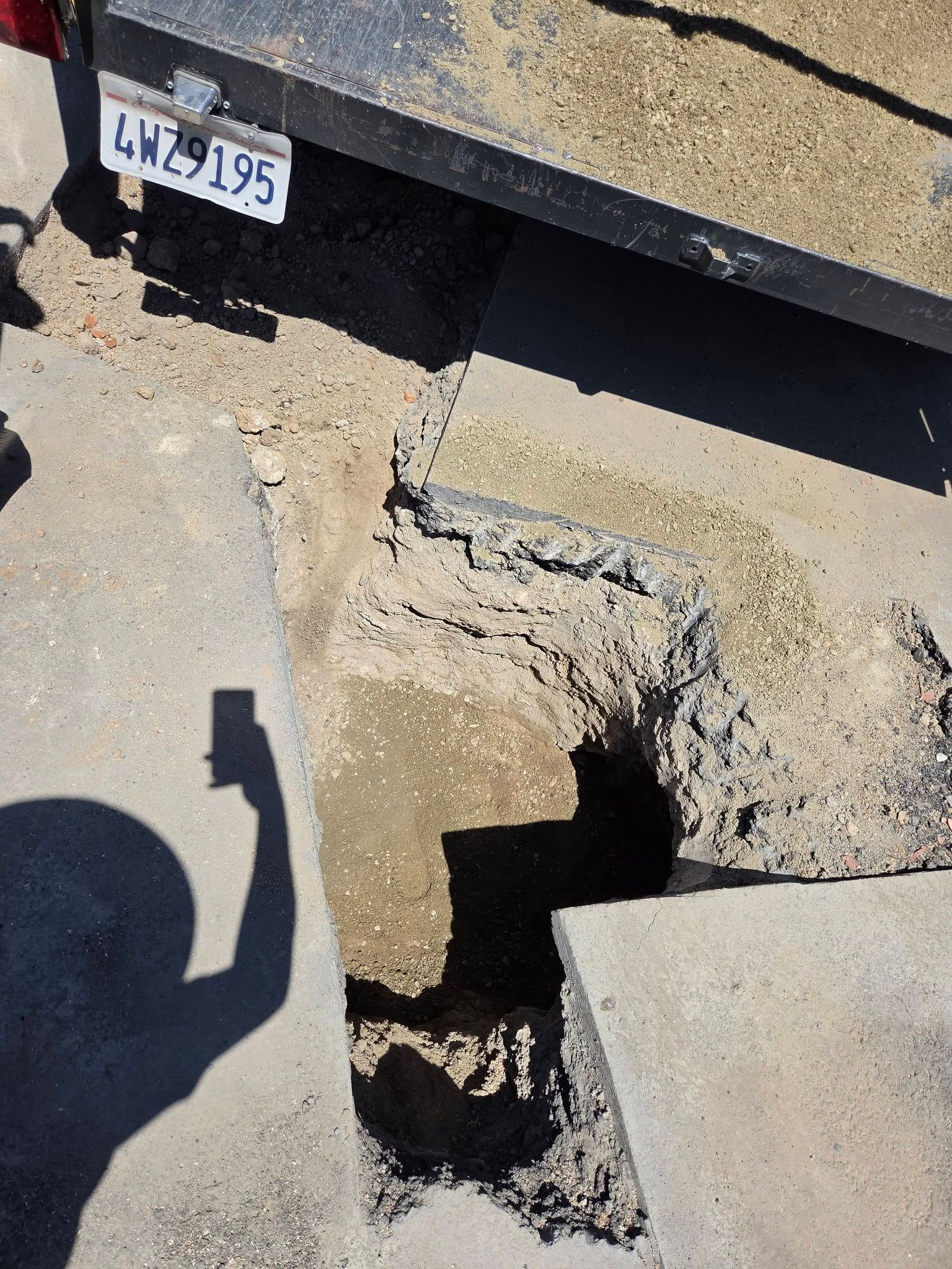 Damaged asphalt and a pothole near a vehicle bumper and license plate on a sunlit street.