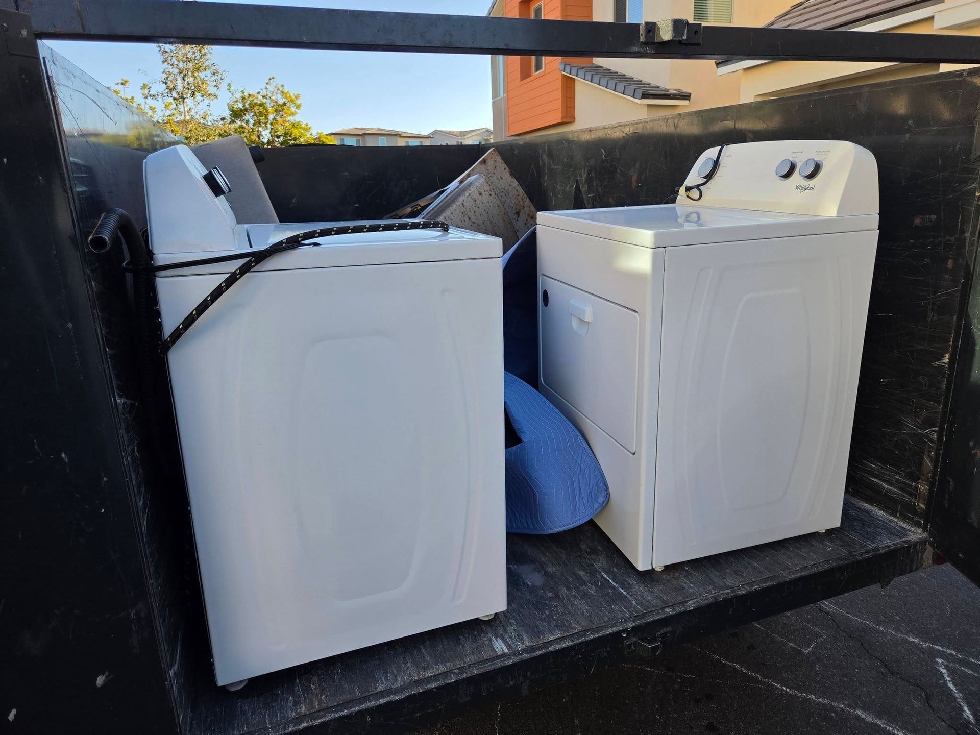 Two white laundry machines in a black truck bed, one washer with its lid open.