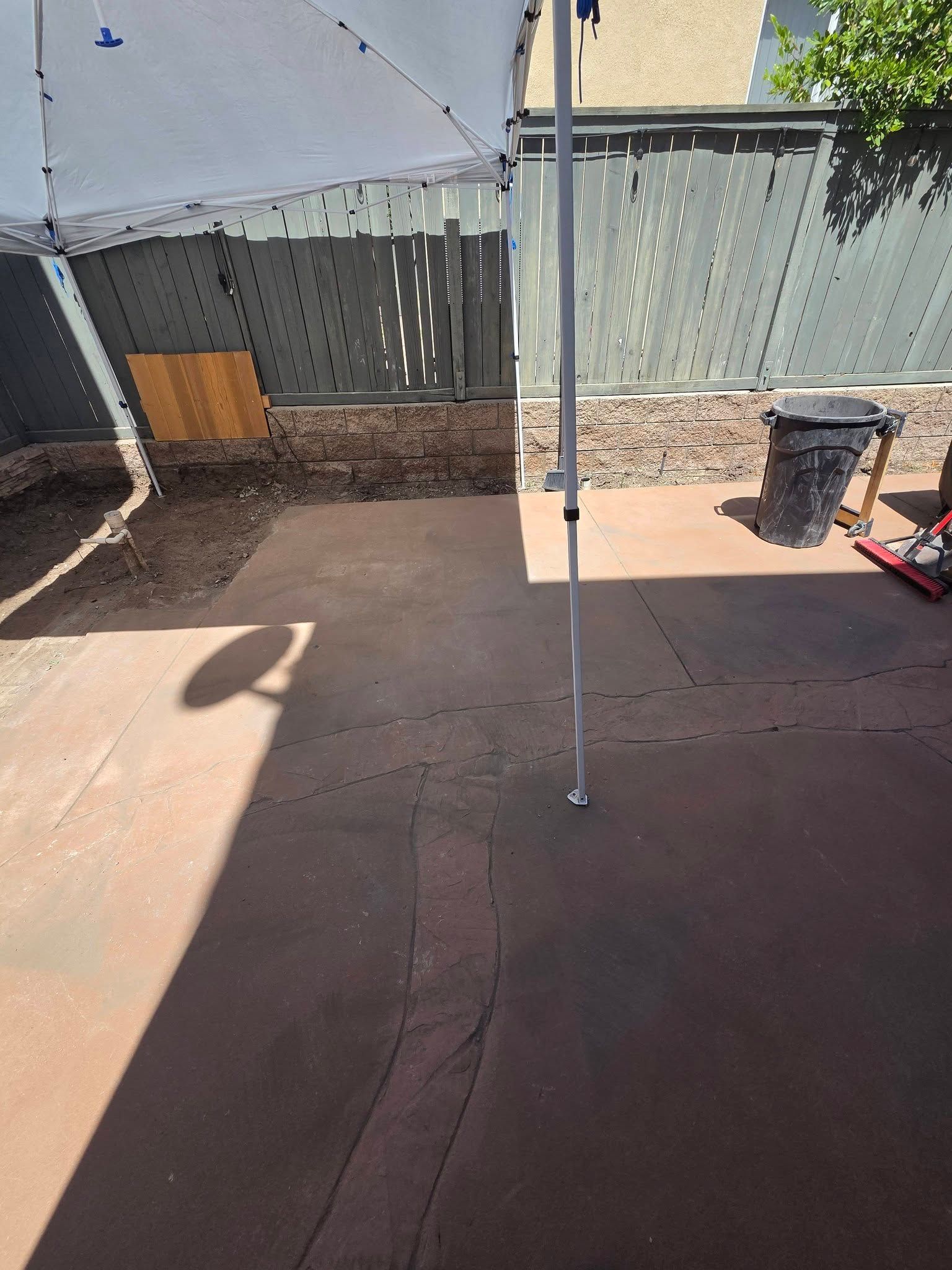Backyard patio with a white canopy pole, red and black mats, and a green fence in bright sunlight