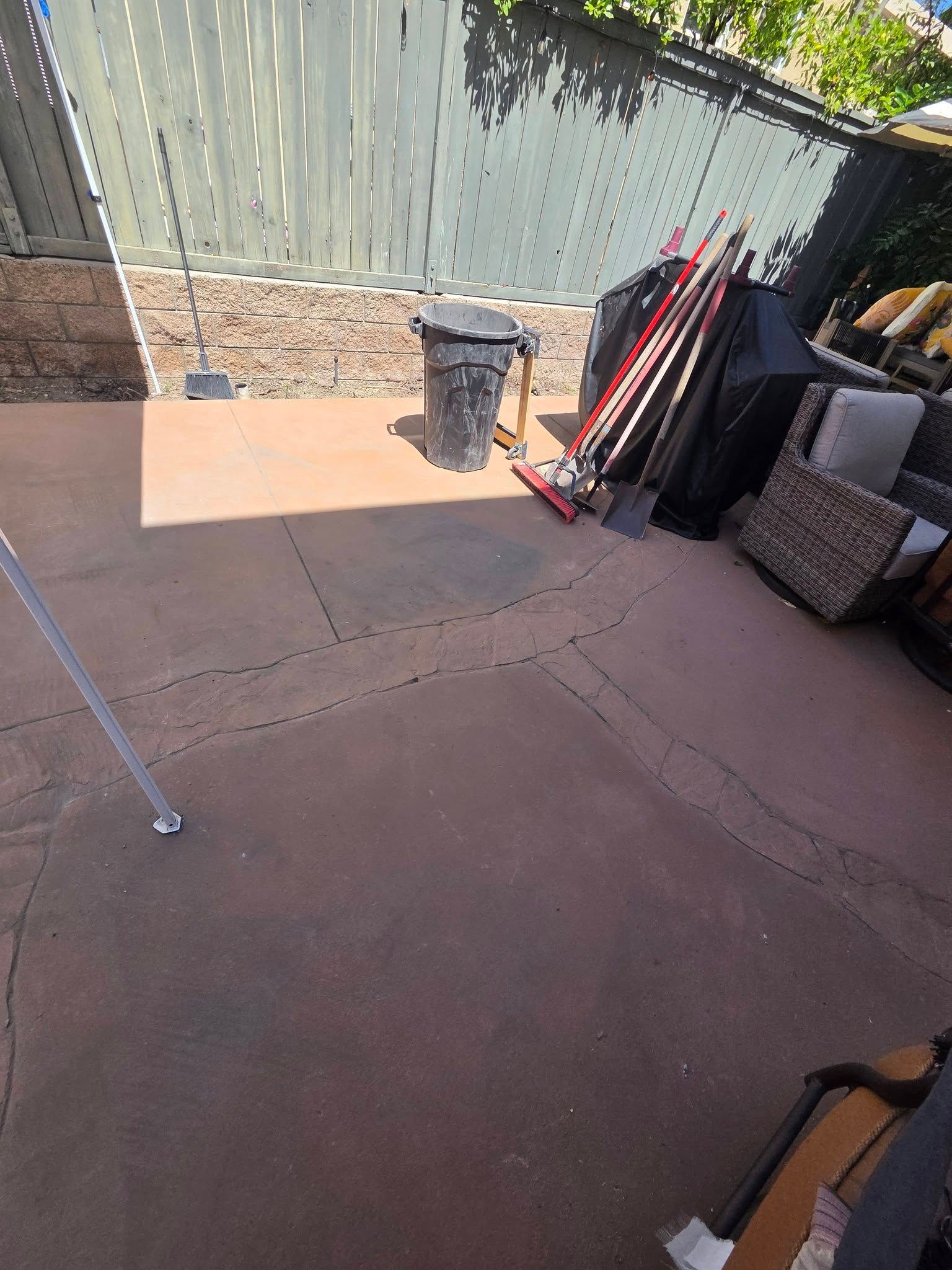 Back patio with concrete ground, green fence, stacked outdoor items, and a trash bin in sunlight
