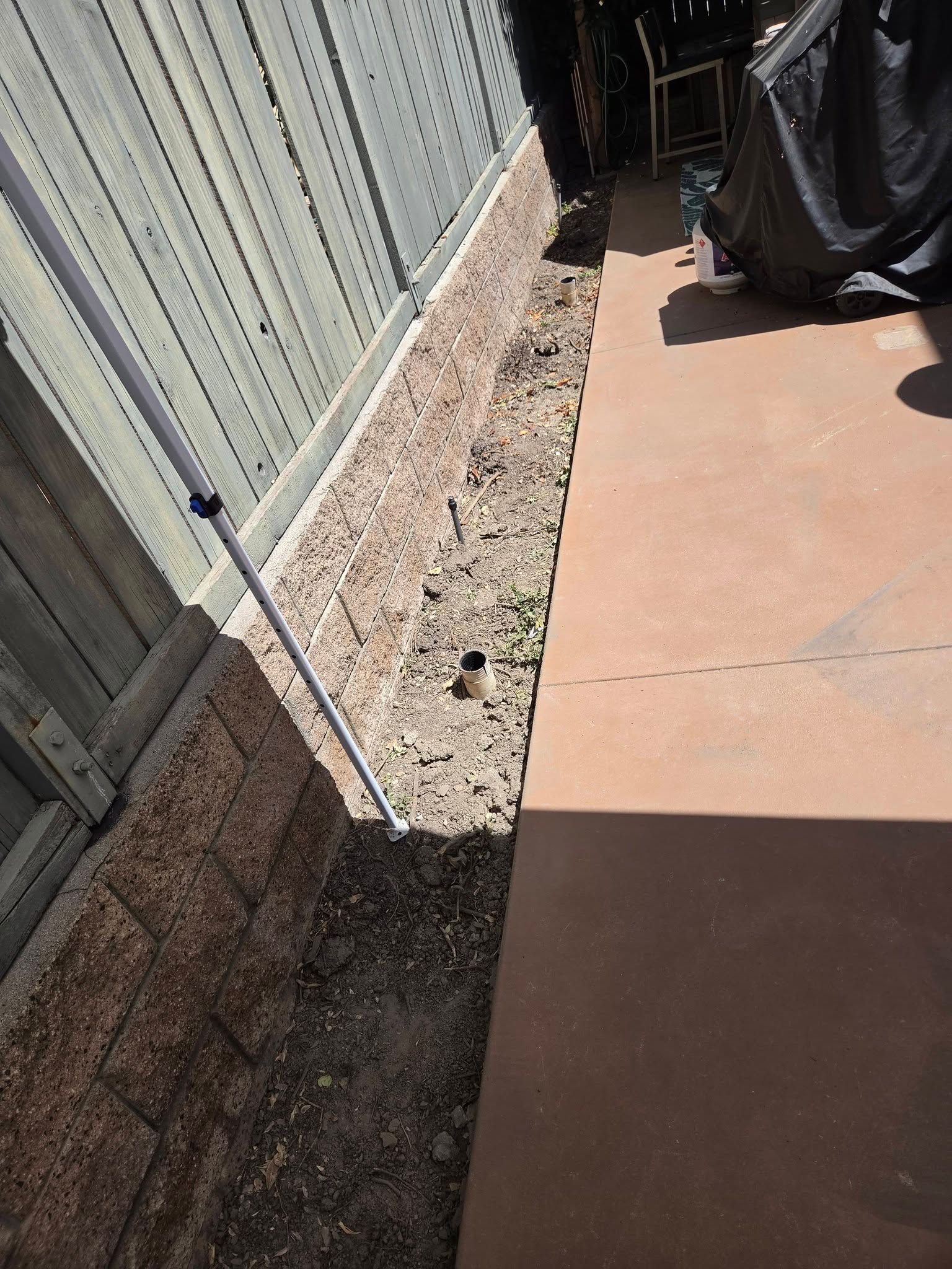 Narrow gravel strip between a metal fence and concrete patio beside a covered object.
