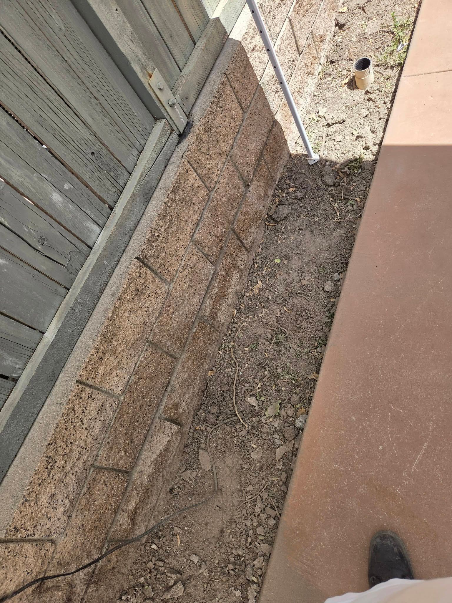 Narrow gravel strip beside a wooden fence and concrete walkway in bright daylight.