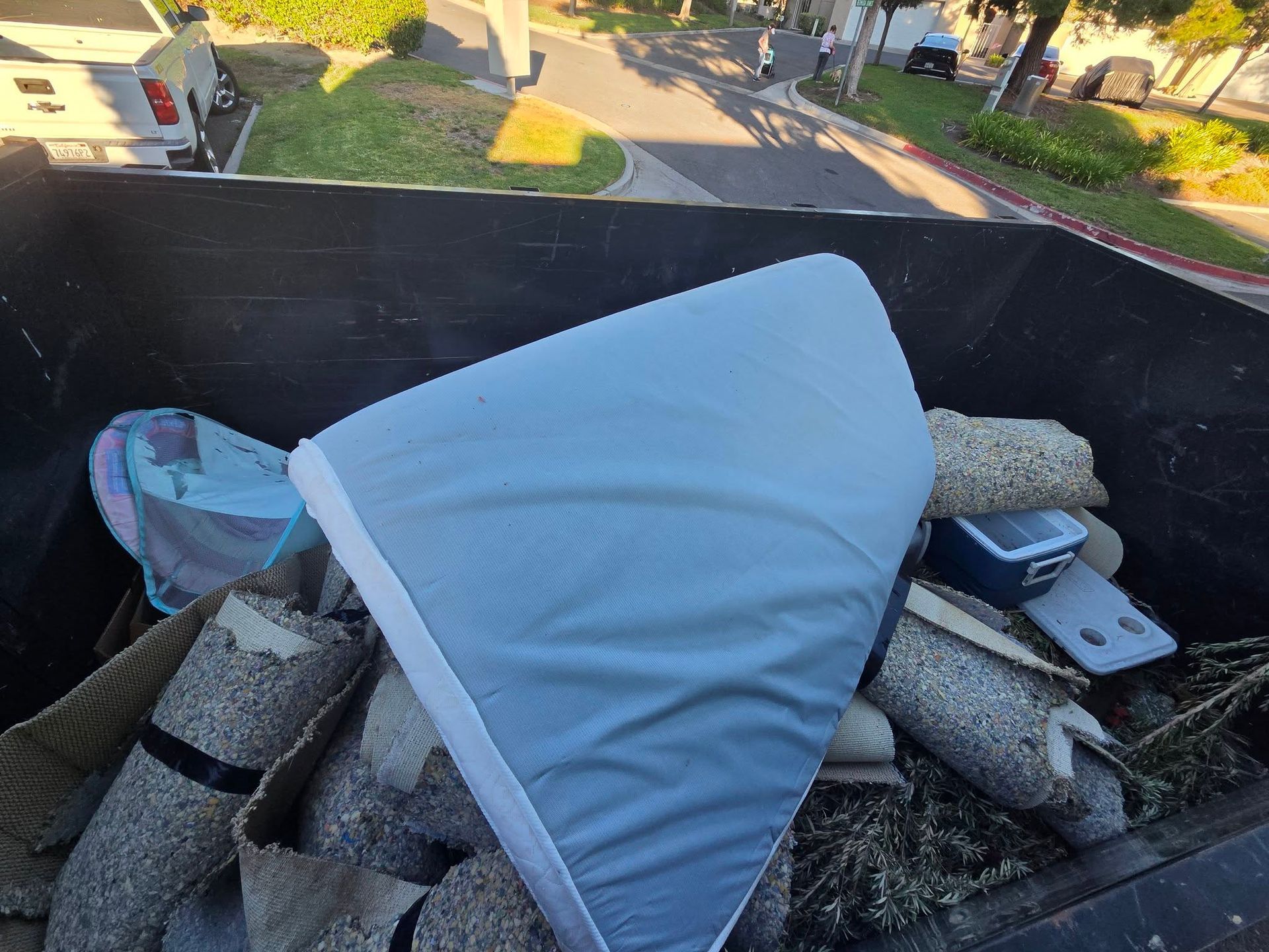 White mattress or foam on a rooftop beside debris and equipment, with a street and houses in the background