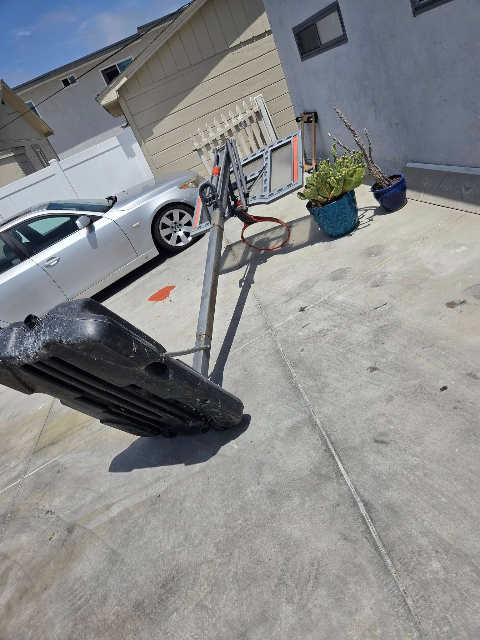 Black trash bin tipped over on a driveway beside a parked silver car and a house with potted plants.