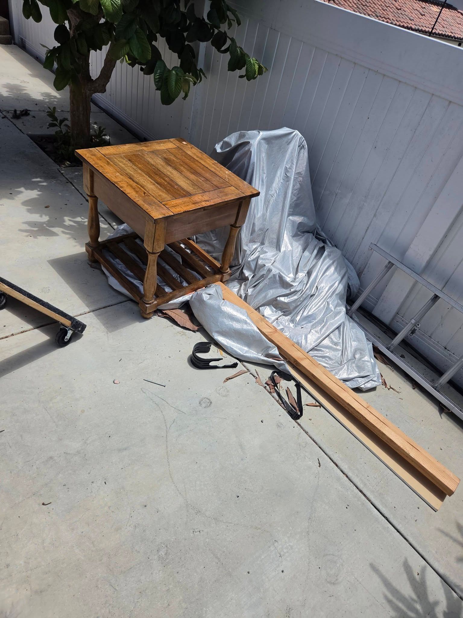 Wooden table and lumber beside a silver tarp on a concrete patio next to a fence