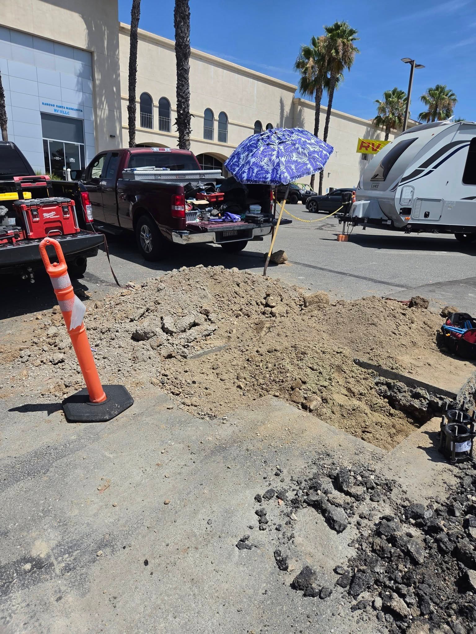 Construction trench beside parked trucks and cones in a sunny parking lot, with palm trees and buildings nearby