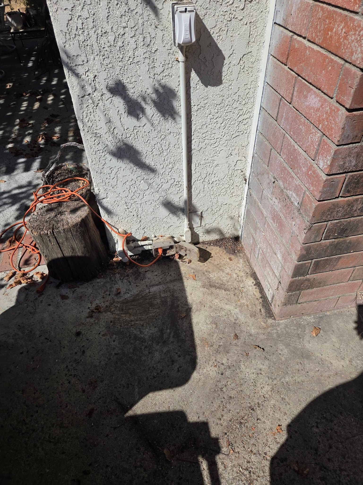 Concrete walkway beside a stucco and brick wall, with a small utility box and tree shadows.