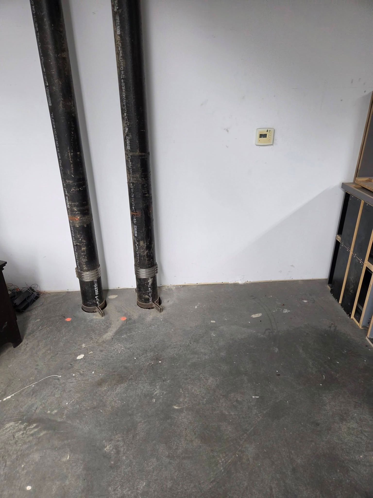 Basement wall with exposed black pipes, gray floor, and a small electrical outlet.