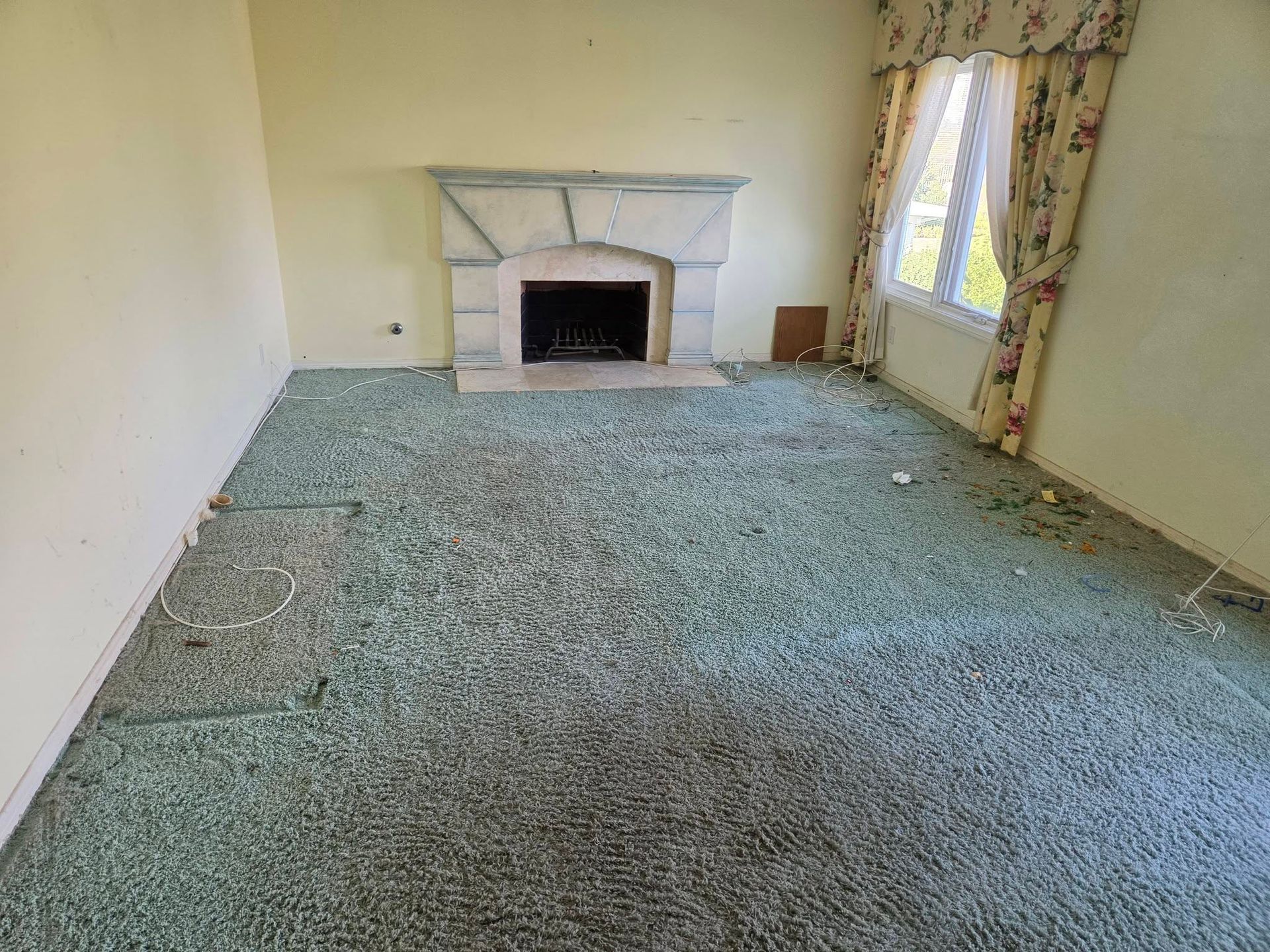 Empty living room with green carpet, fireplace, and window with floral curtains