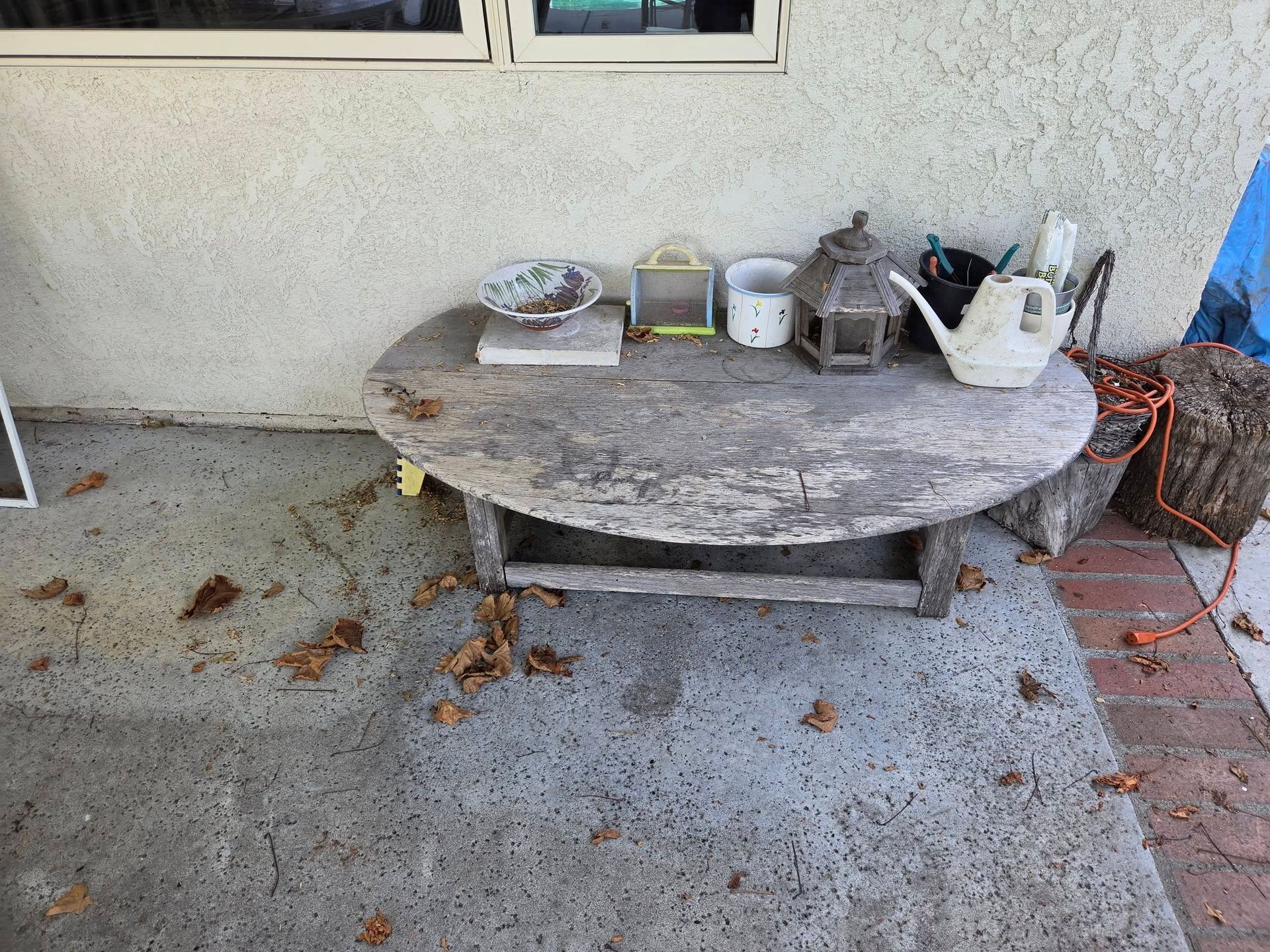 Weathered round outdoor table with potted plants and gardening items on a concrete patio beside a wall