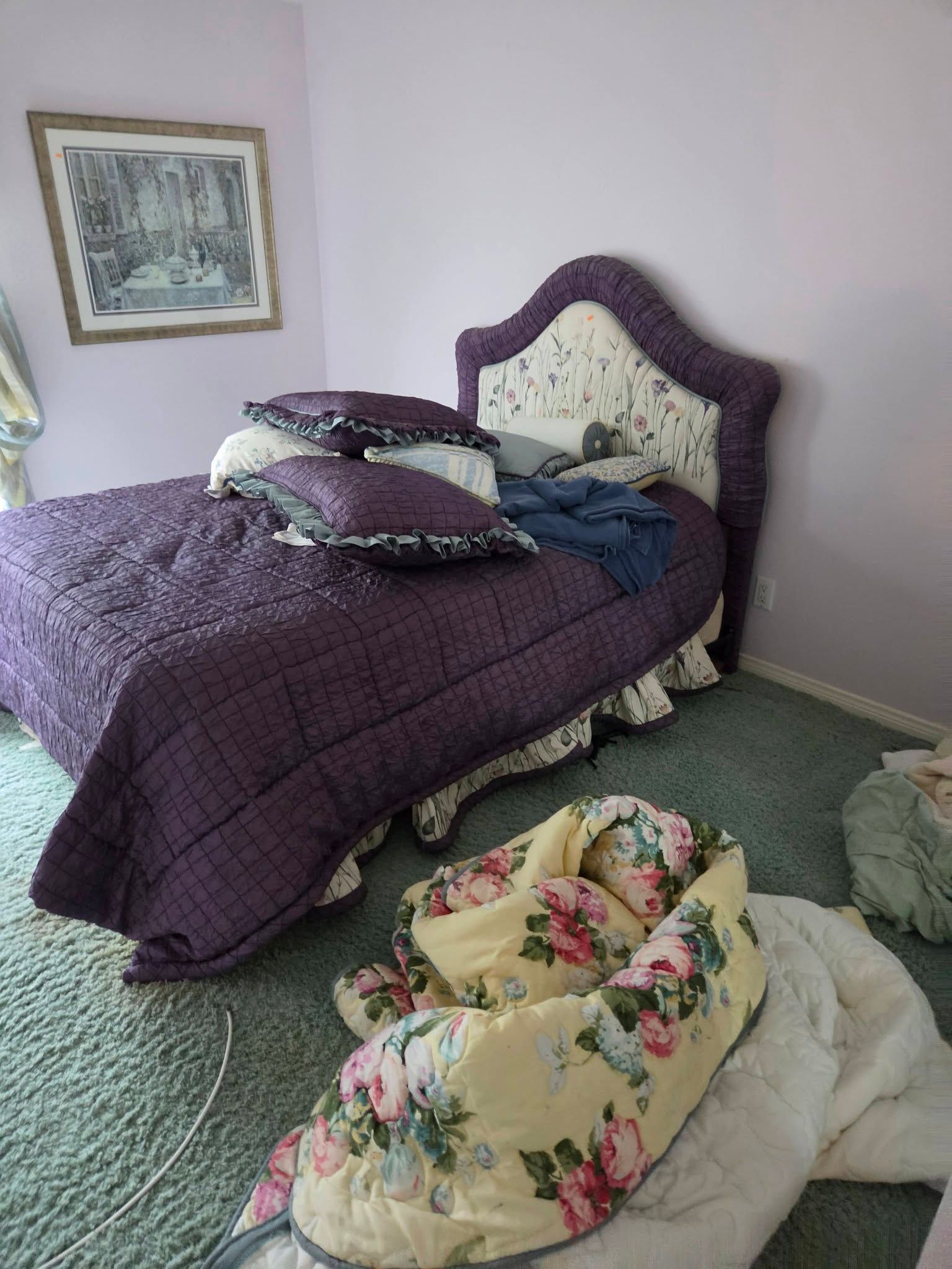 Bedroom with purple bedspread and floral cushions on the floor, green carpet, and framed wall art.