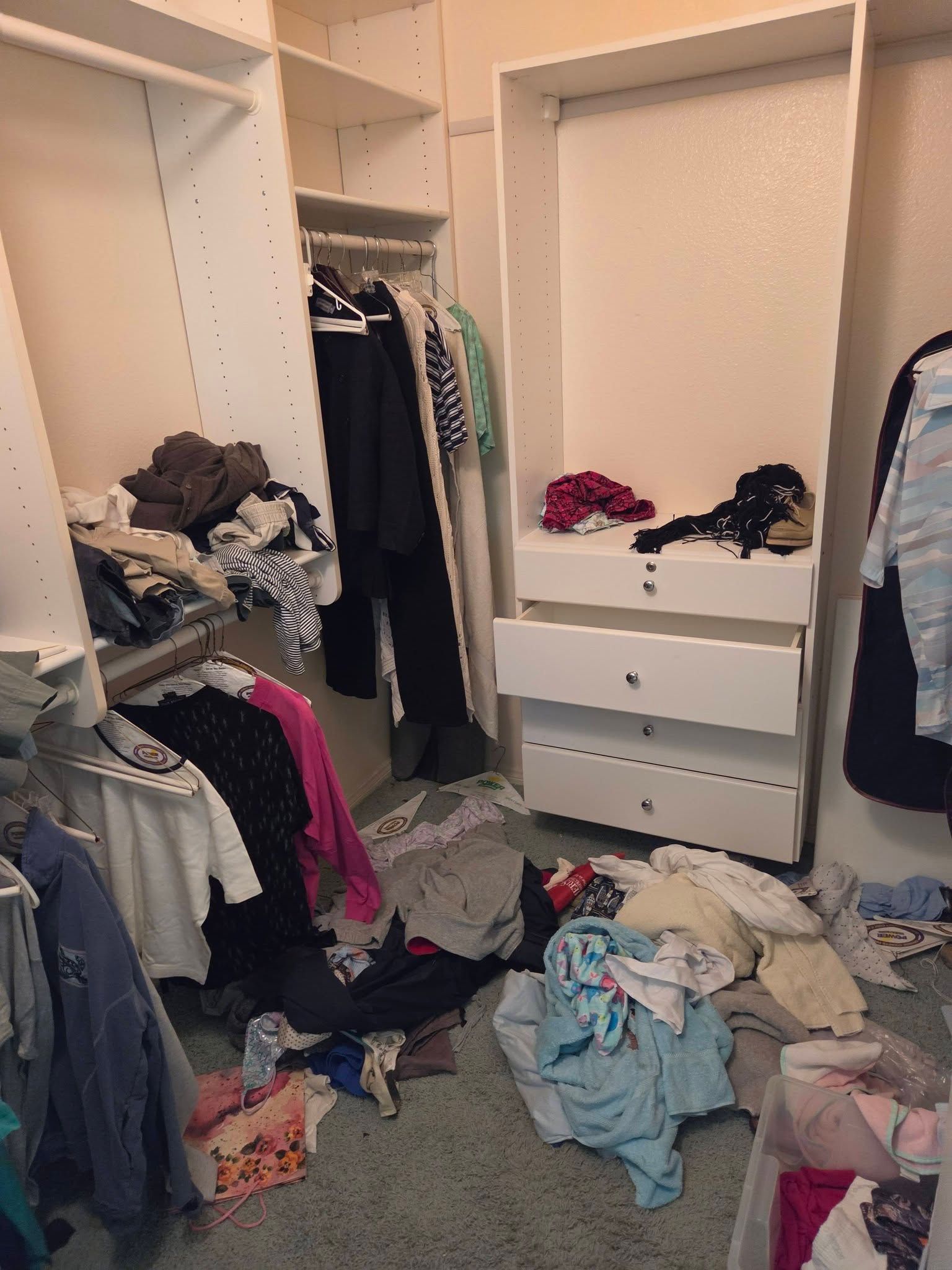 Cluttered walk-in closet with clothes piled on the floor and hanging from shelves and rods.