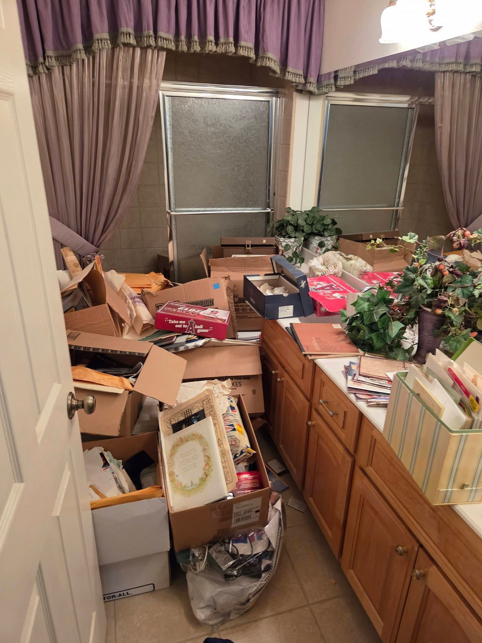 Cluttered room with boxes and bags piled on the floor beside cabinets and windows with purple curtains