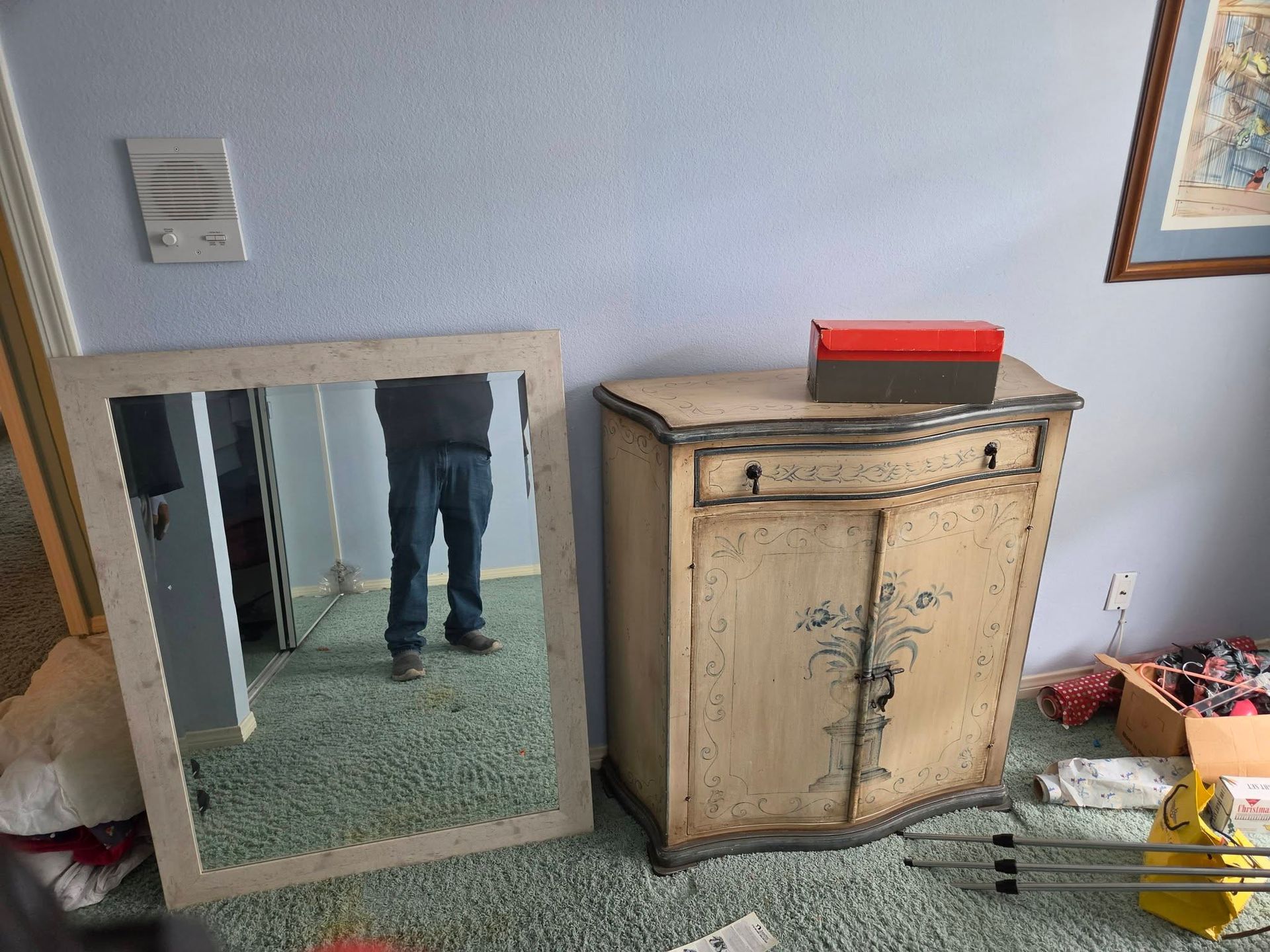 Mirror leaning on wall beside a beige cabinet in a sparse room, with a person’s legs reflected in the mirror.