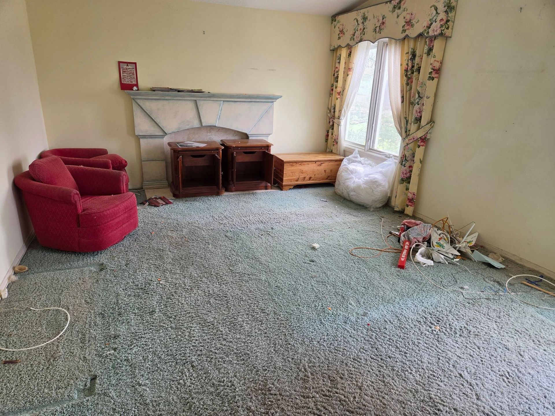 Empty living room with green carpet, red chair, wooden shelf, and floral curtains by the window.