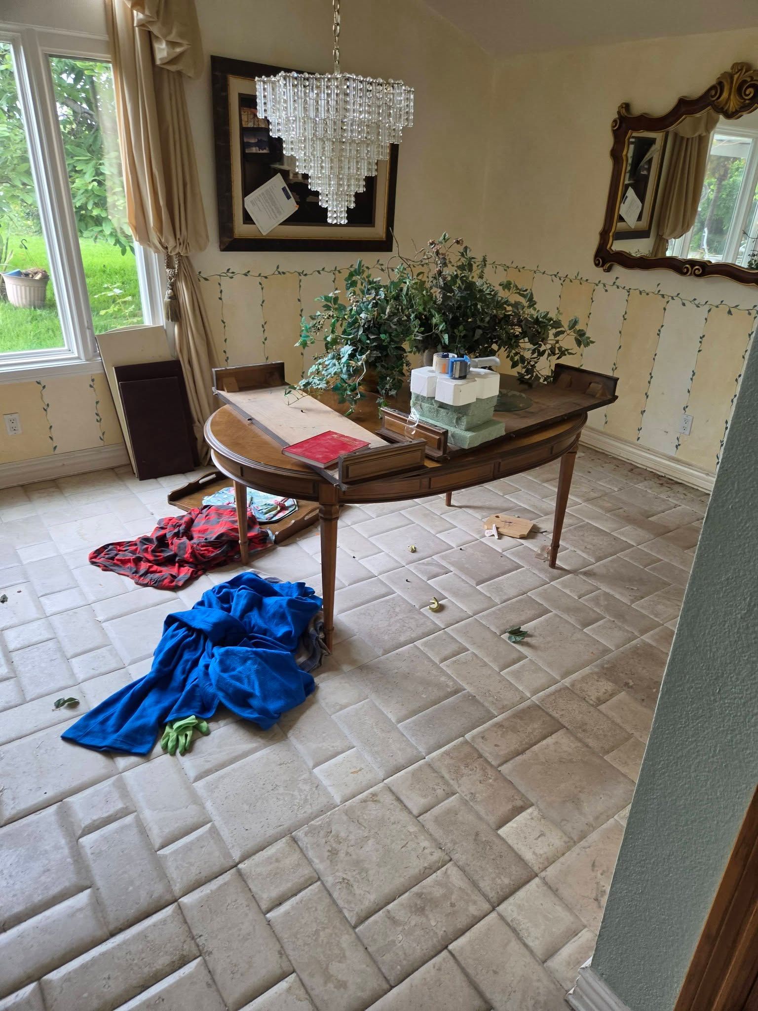 Cluttered dining room with a wooden table, mirror, flowers, and clothes on the floor.
