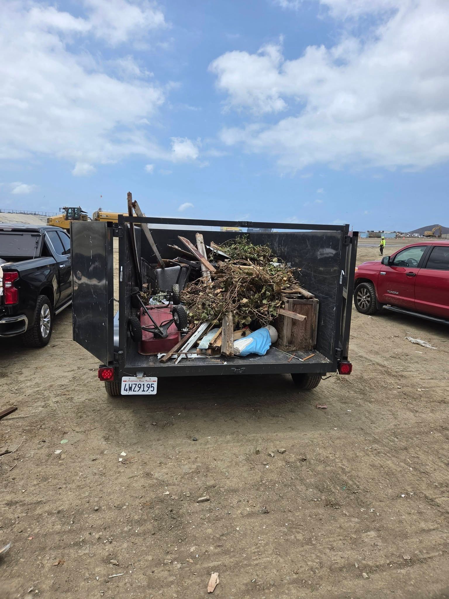 Trash-filled utility trailer in a gravel lot under a partly cloudy sky, with cars on either side