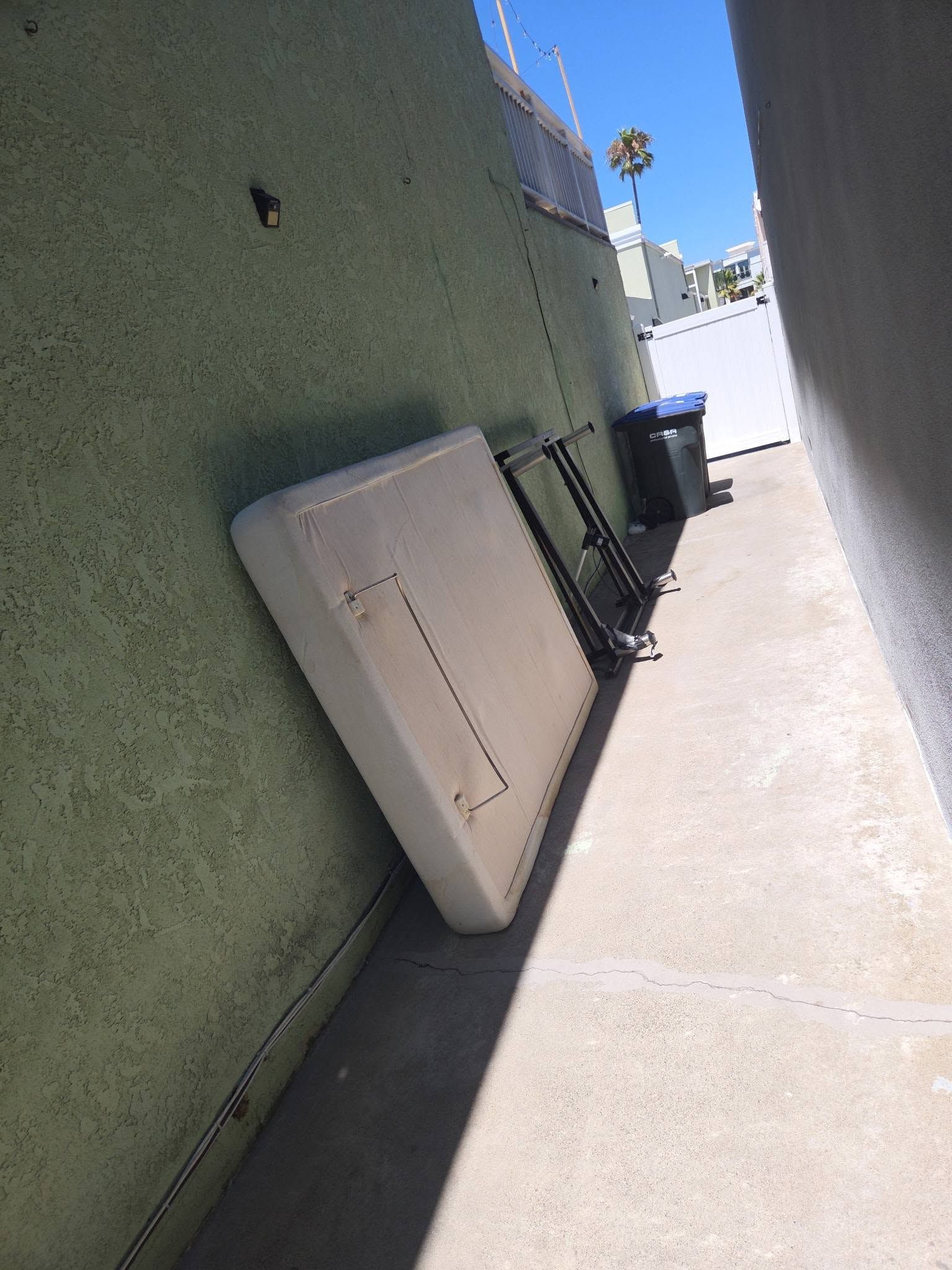 Narrow outdoor alley with green and white walls, a leaning mattress, trash bin, and bright sunlight.