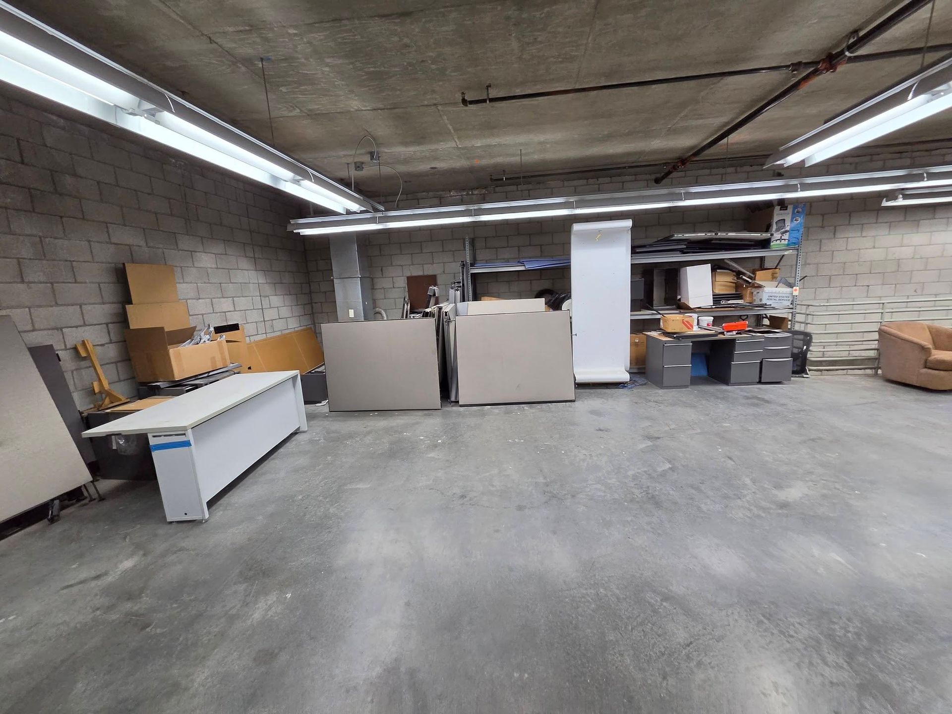 Empty industrial basement with concrete floor, fluorescent lights, and scattered storage cabinets and boxes