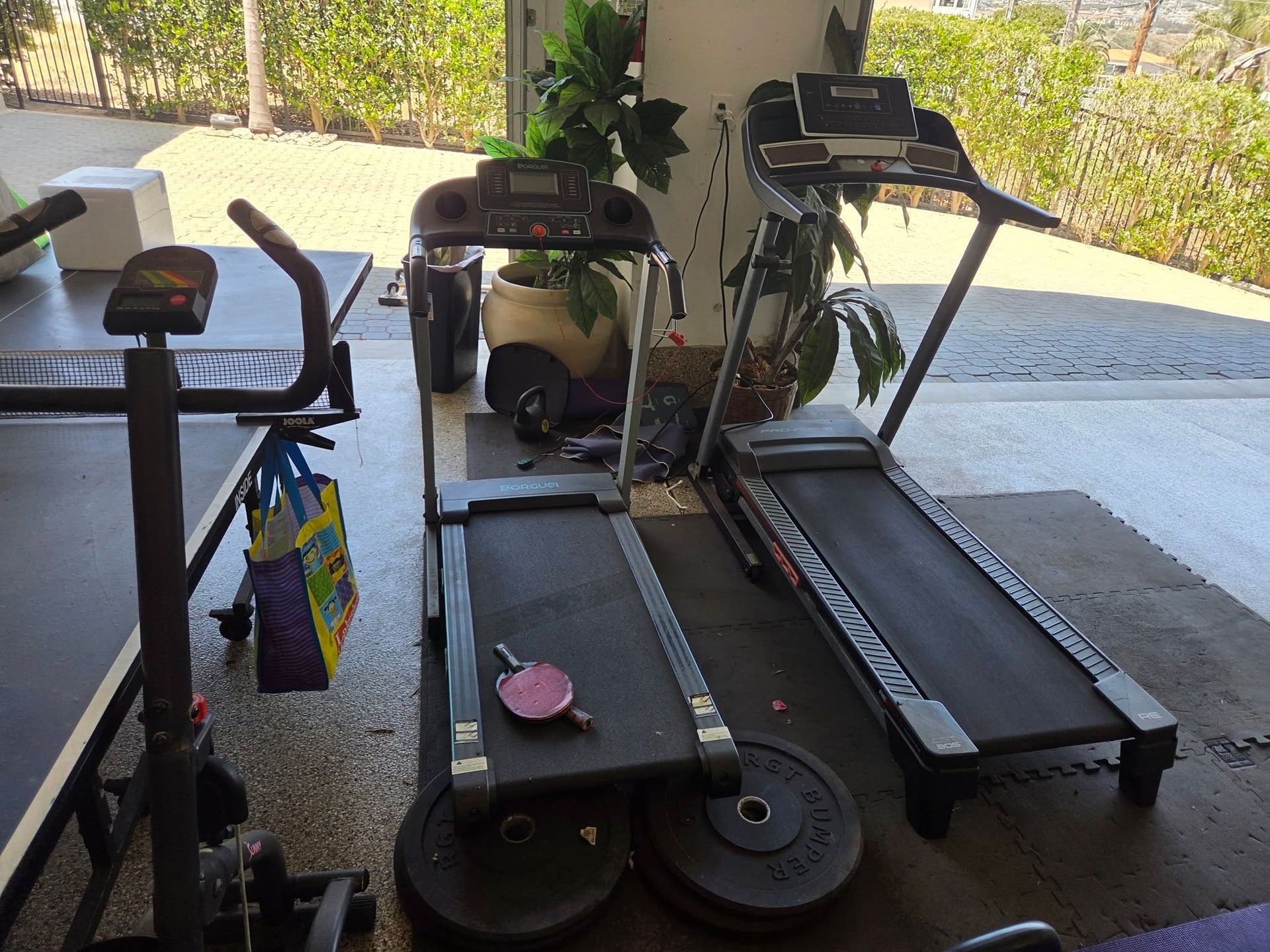 Three treadmills and an exercise bike in a covered garage area with plants and a driveway beyond.