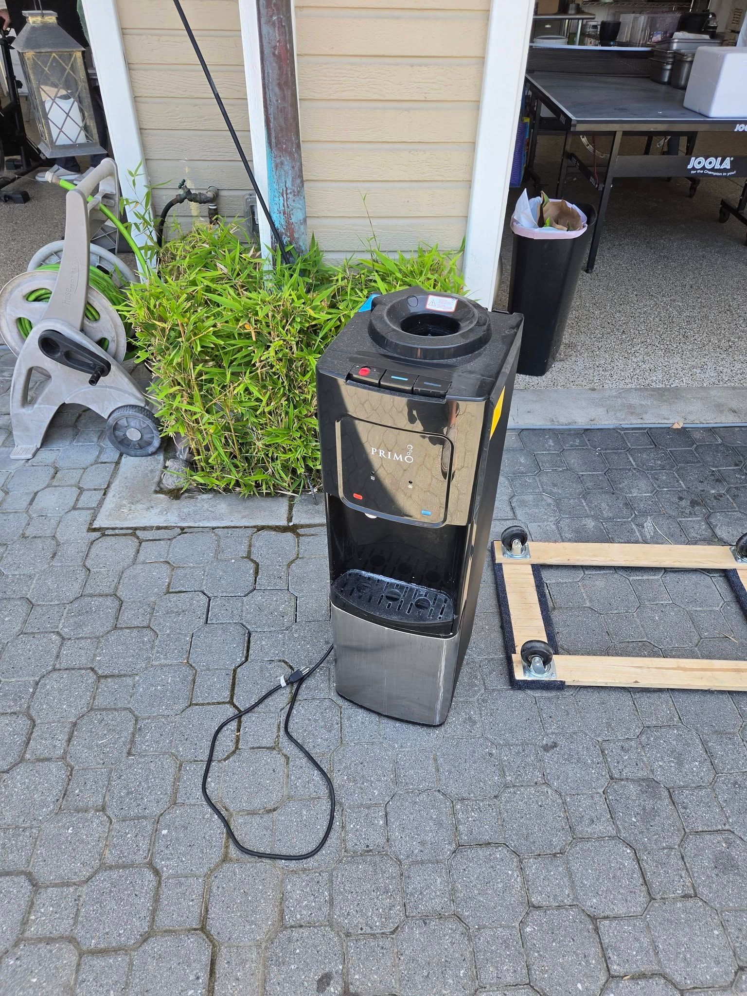 Black damaged speaker or machine on pavement outside a building with plants and scattered cords