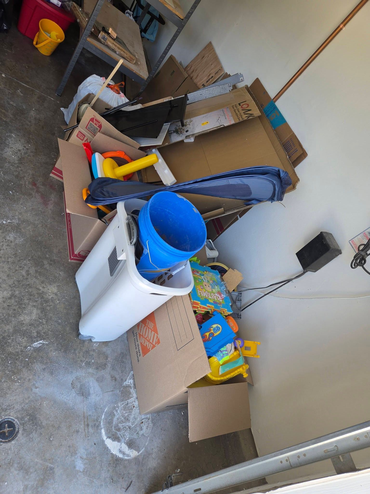 Cardboard boxes and assorted tools stored in a narrow outdoor service area with a white bin and blue bucket