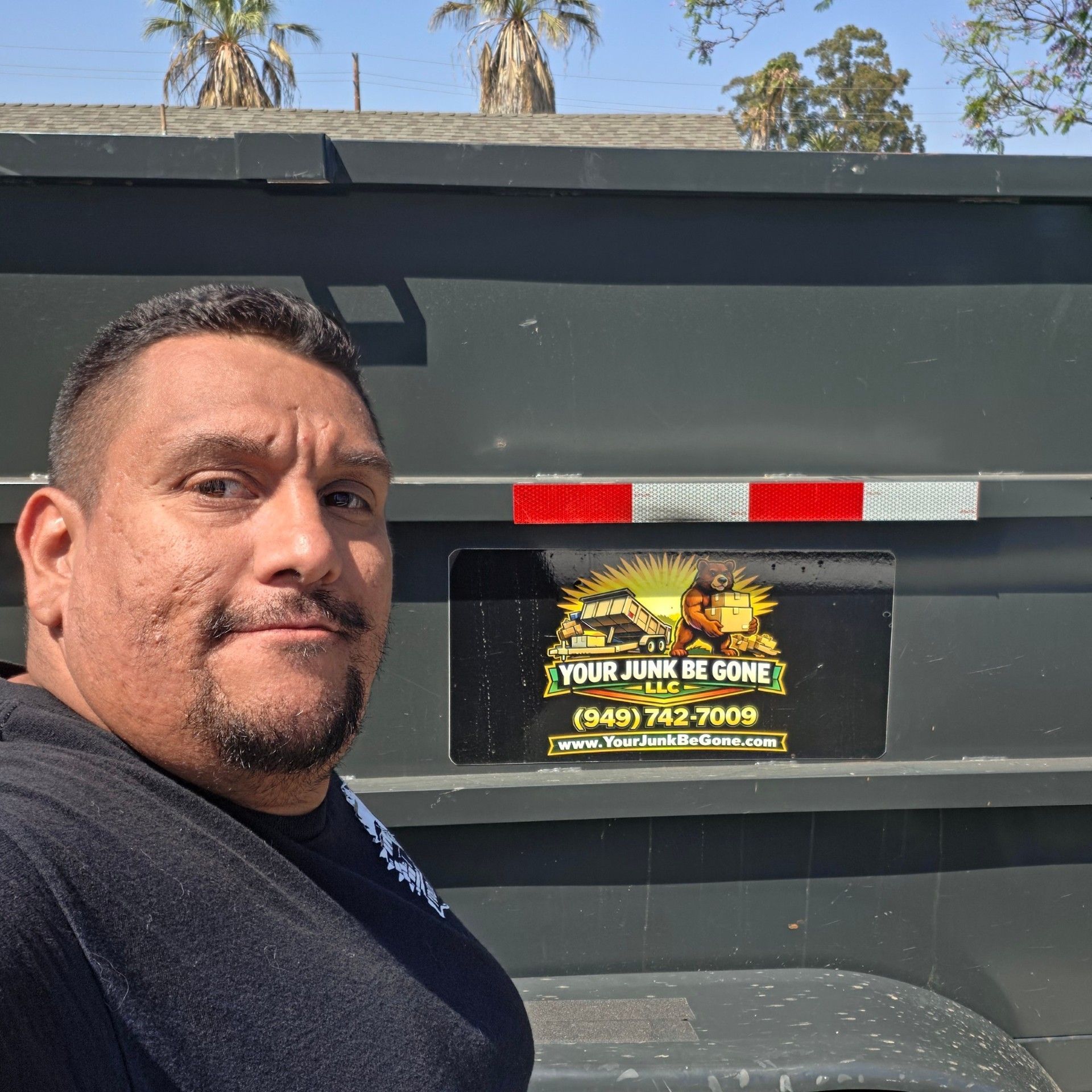 Man standing beside a dark dumpster with a warning sign and red-white reflective strip on the back.